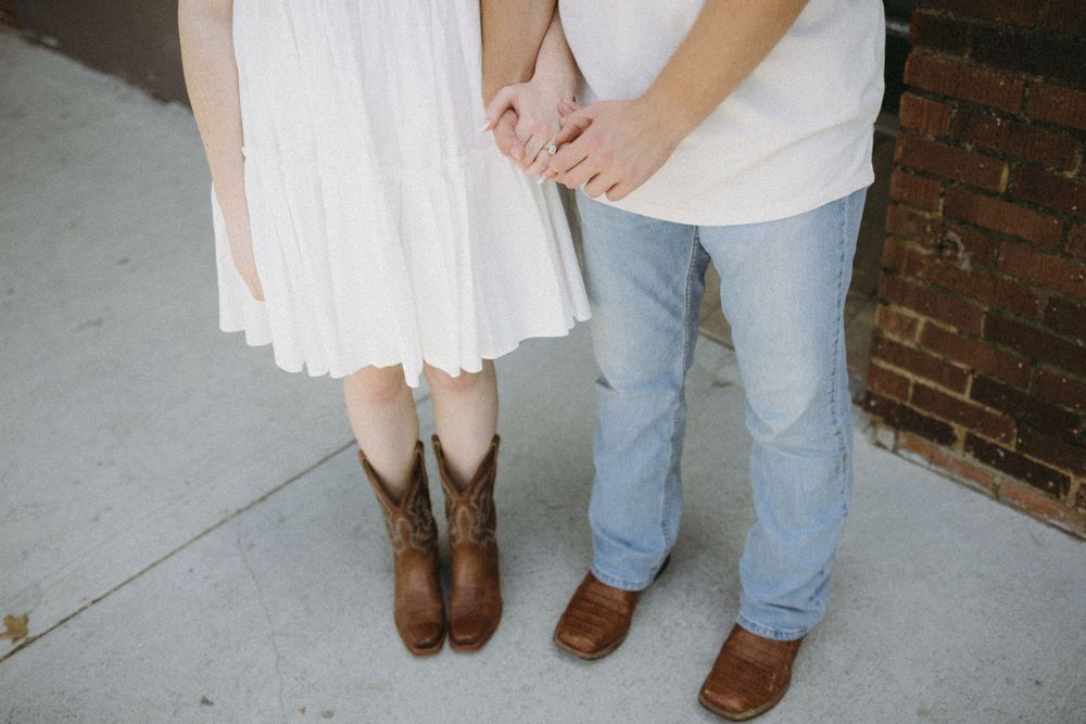 Couple holding hands, woman in white dress with cowboy boots and man in jeans and loafers on concrete sidewalk.