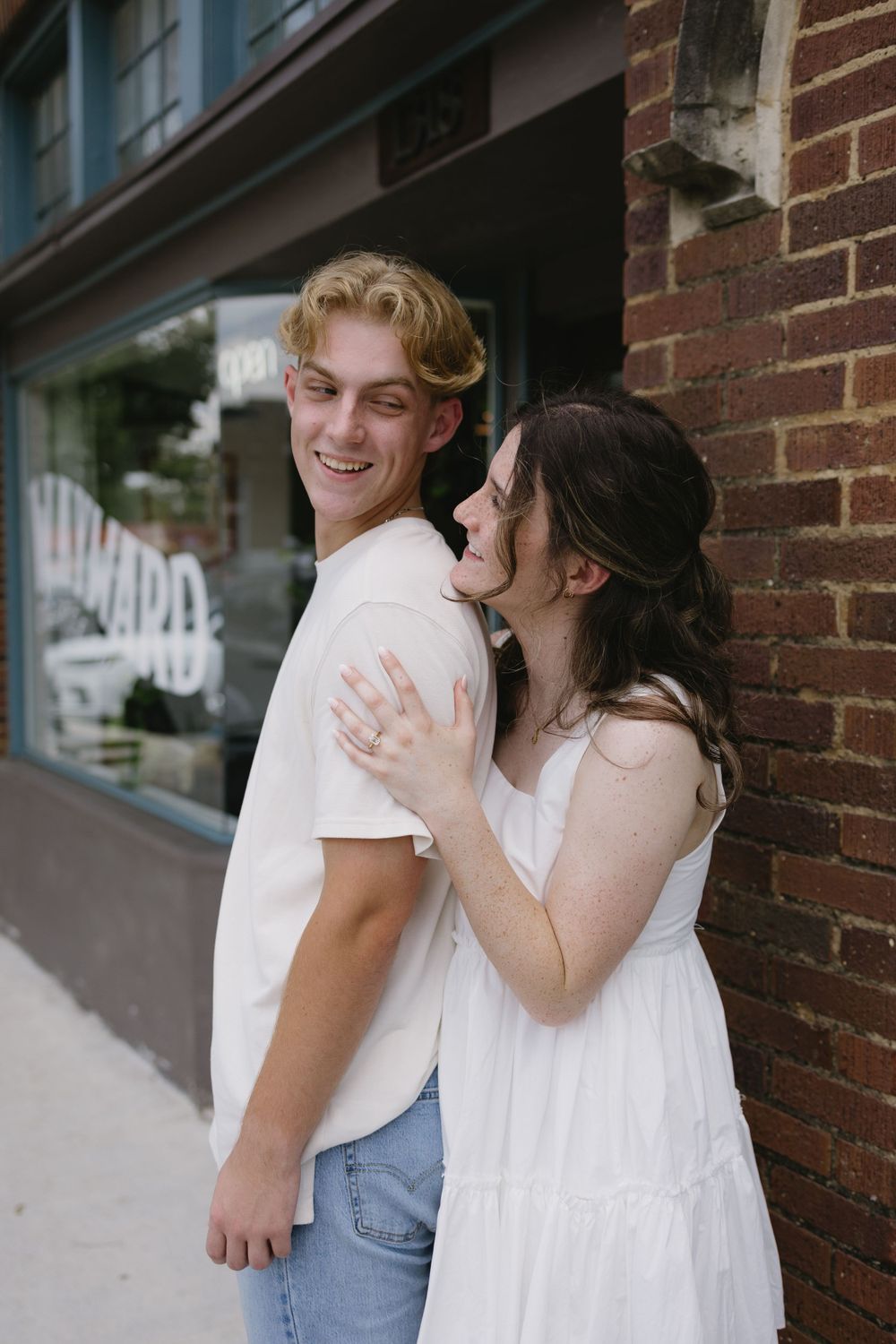 Smiling couple embracing outside a brick building, man in white tee and jeans, woman in white sundress.