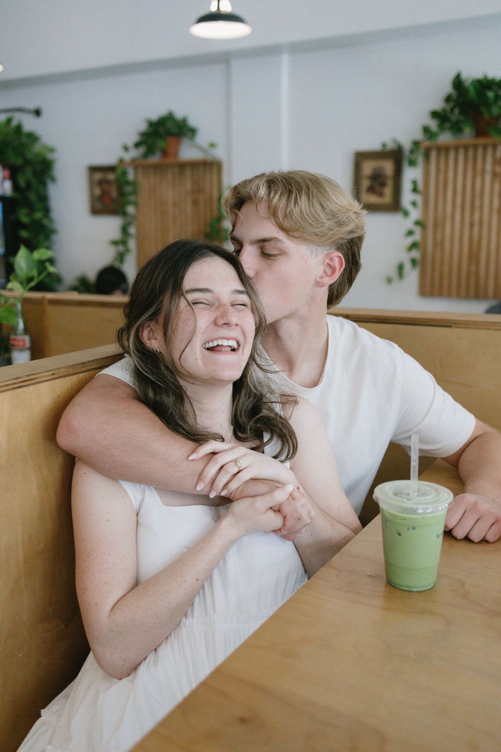 Man kissing woman on the cheek as she laughs in a cafe booth, iced matcha drink on the table.
