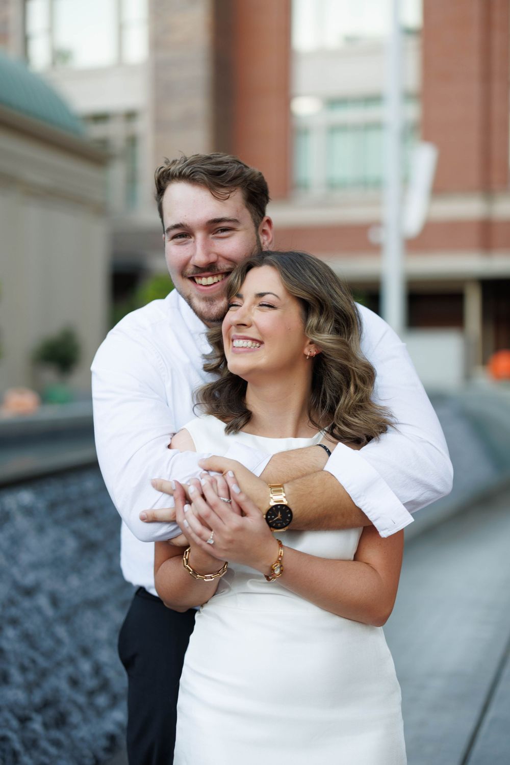 Smiling couple hugging near a fountain, man in white shirt wrapping arms around woman in white dress.