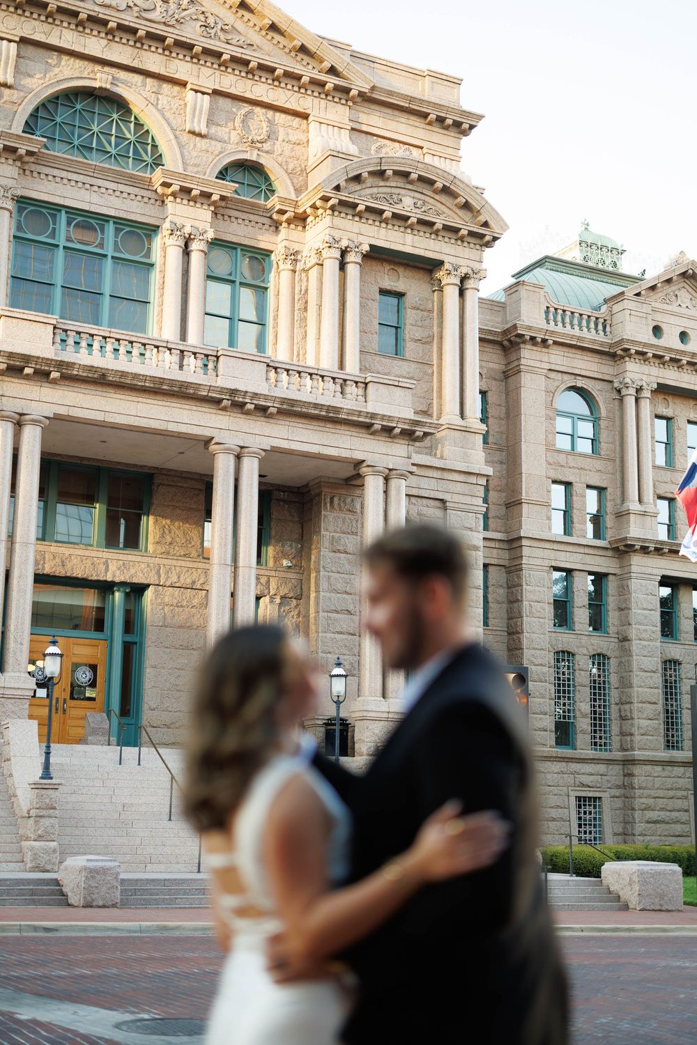Couple sharing a romantic embrace in front of a grand historic stone courthouse building.