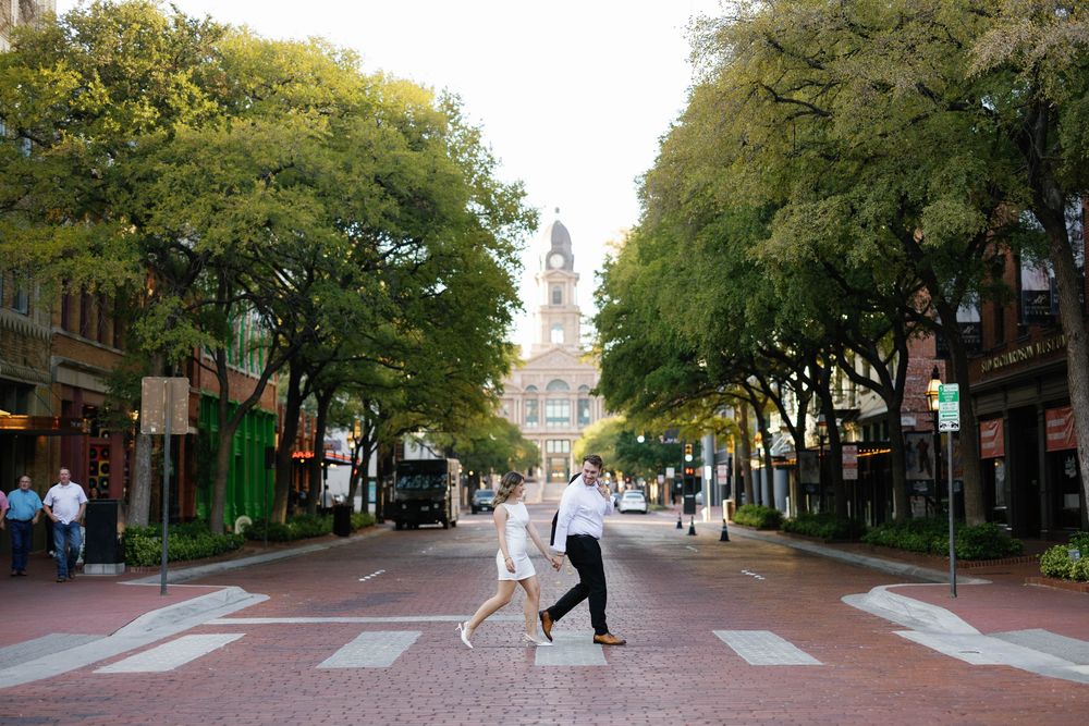 Couple crossing a brick street in downtown area lined with trees and historic buildings.