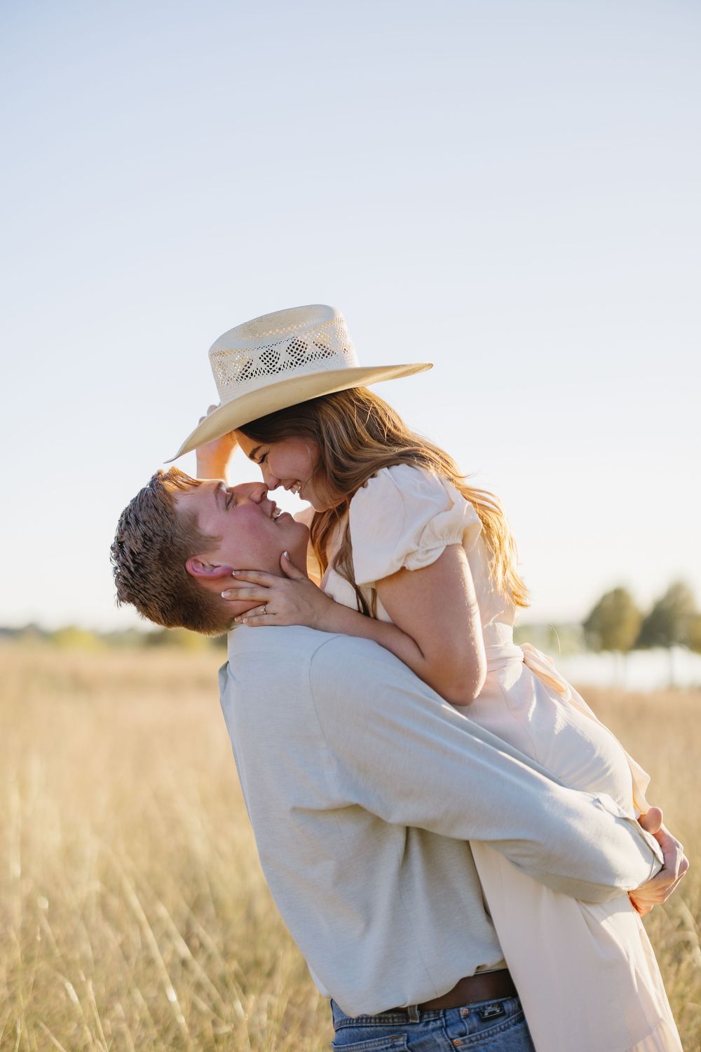 Romantic couple almost kissing as man lifts woman in straw hat in sunlit golden field.