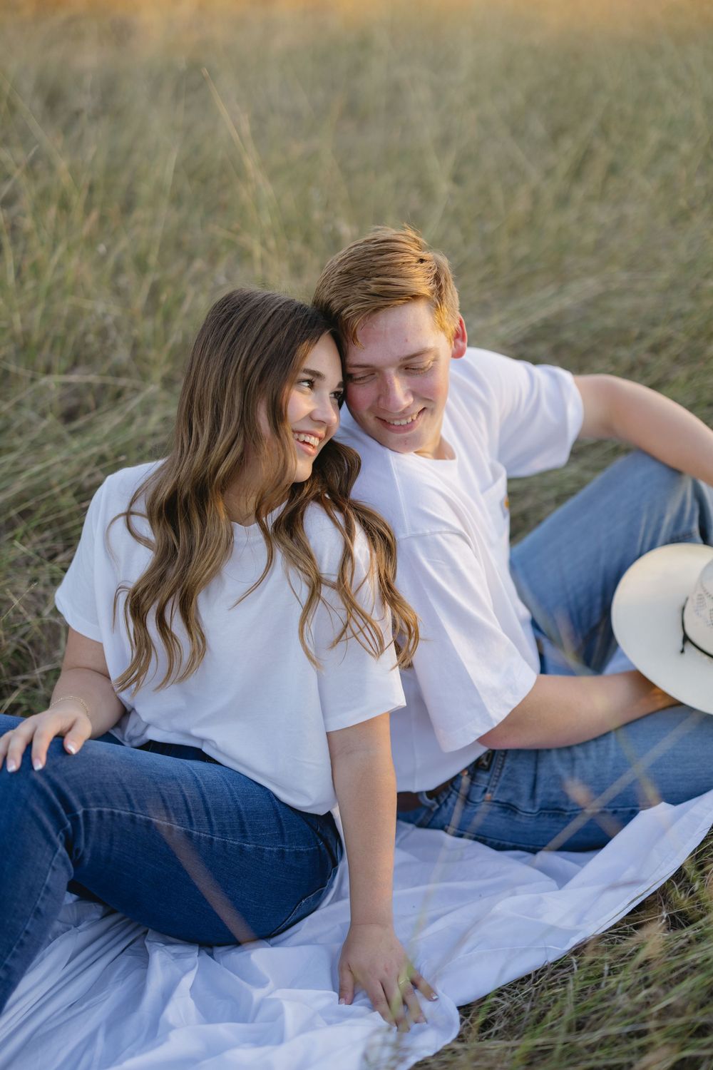 Happy couple sitting together on white blanket in grassy field, both wearing matching white tees and jeans.