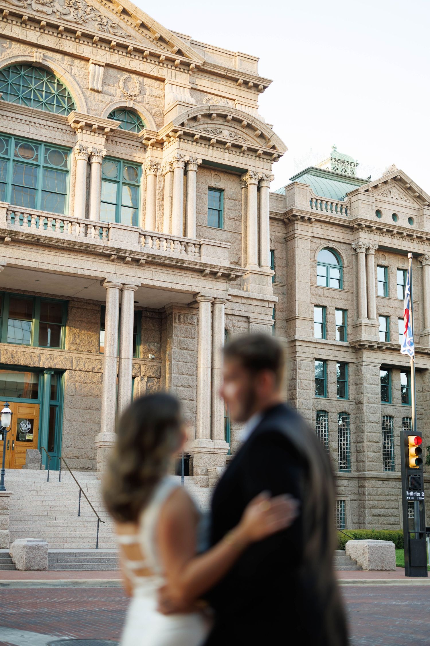 Bride and groom share romantic embrace in front of grand historic stone courthouse building.