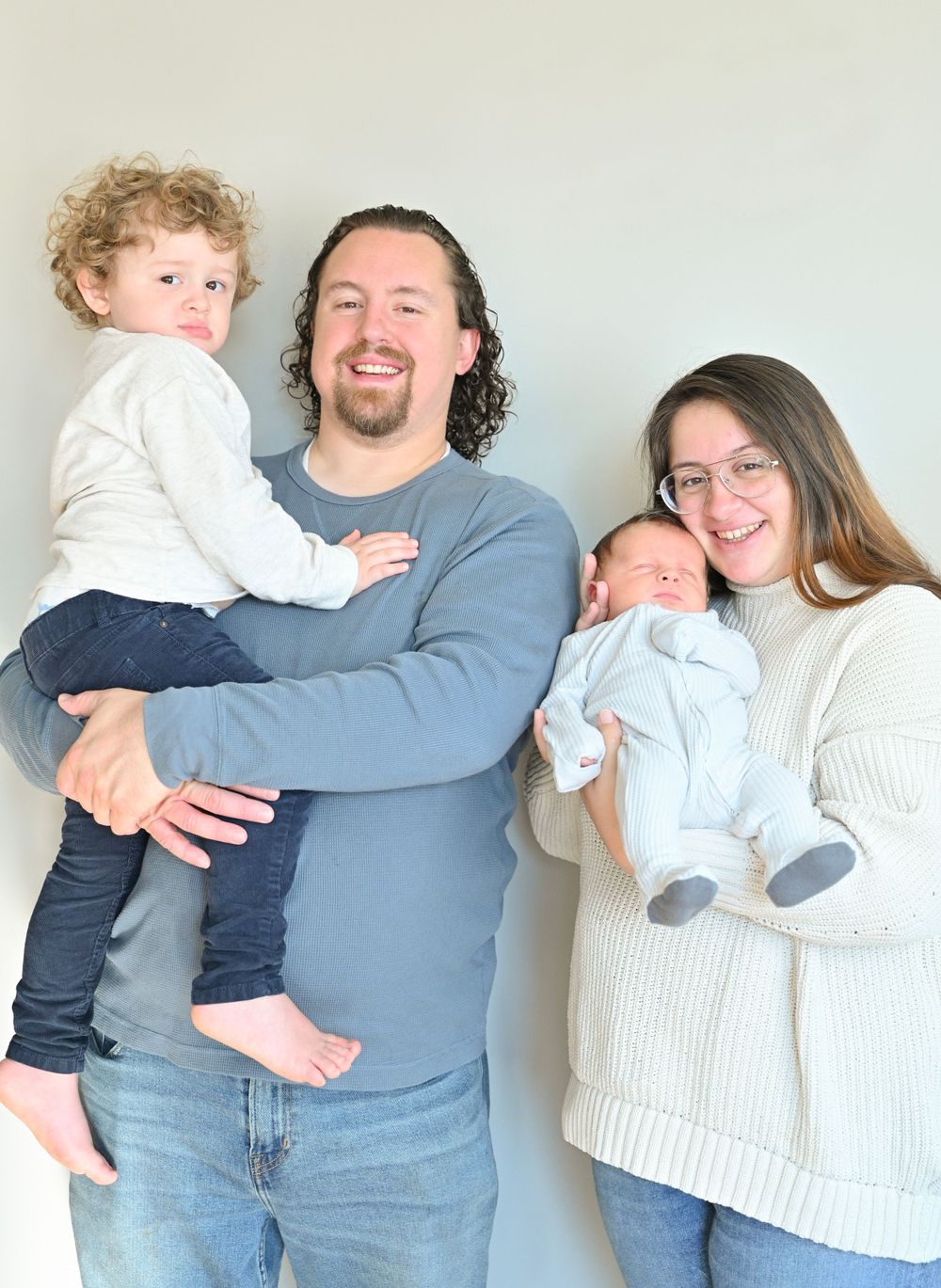A family of four poses together against a white wall wearing casual sweaters.
