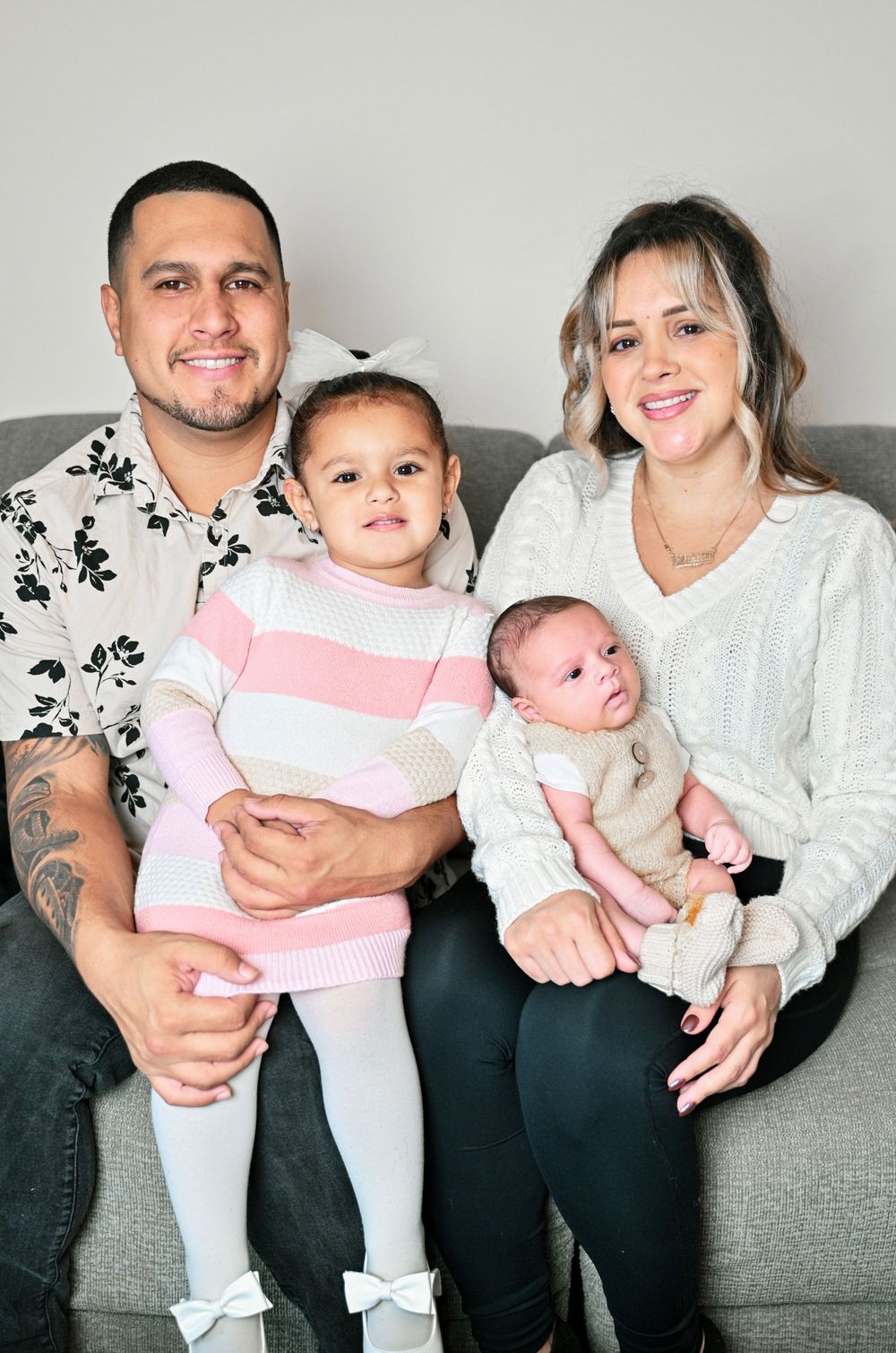 A family sits together on a grey couch with their two young children.
