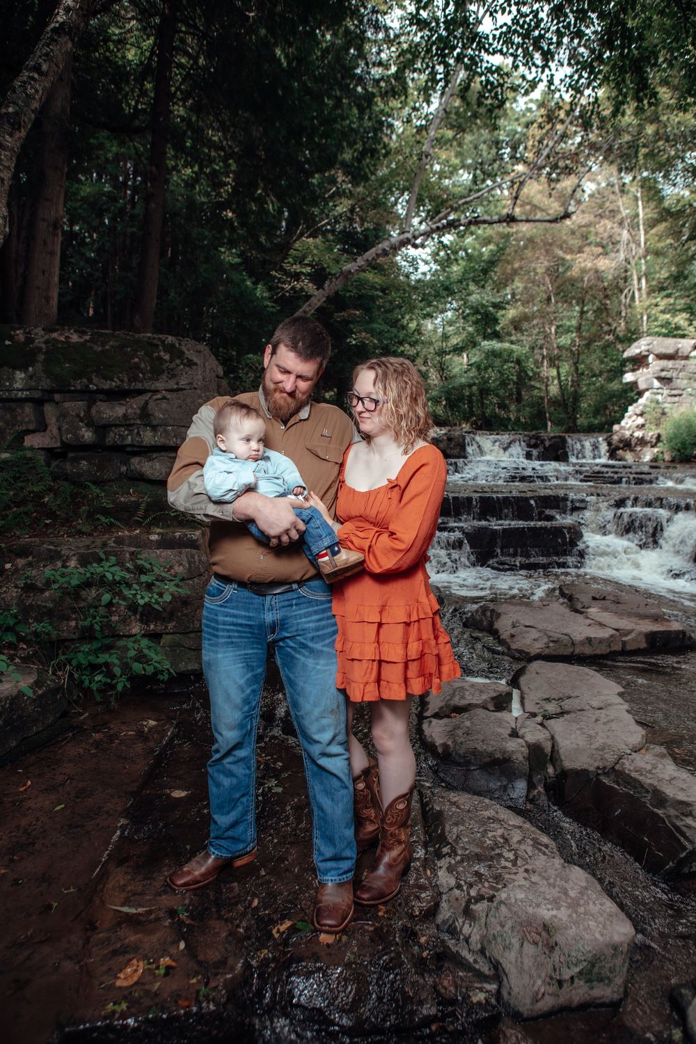 A family stands together on rocky terrain near a cascading waterfall in a wooded setting.