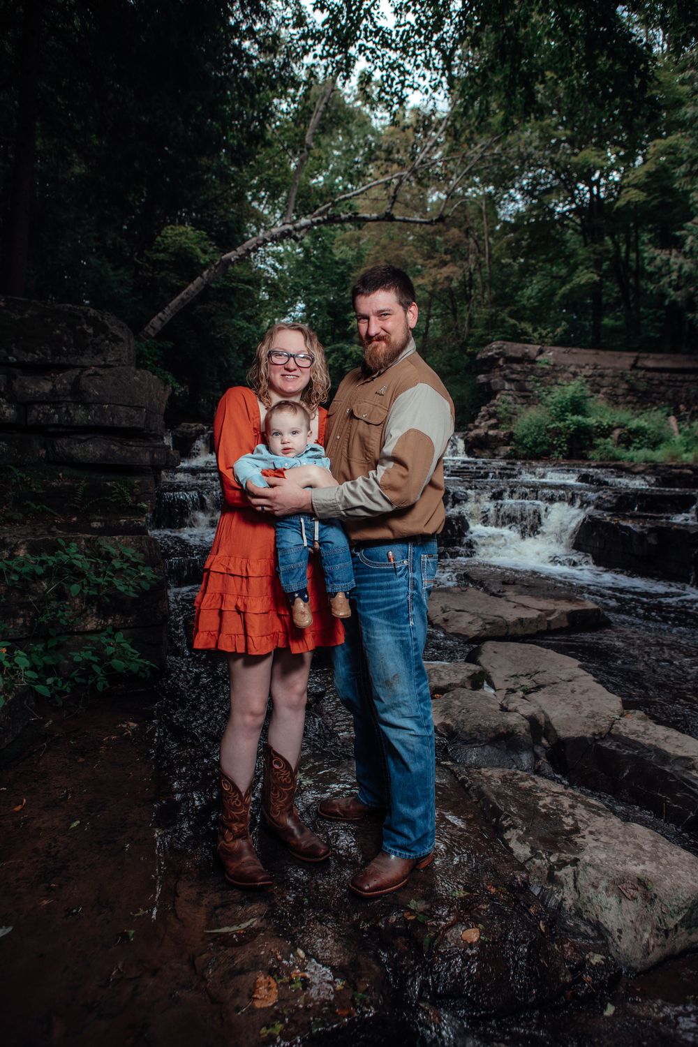 A family poses together near a scenic waterfall on rocky terrain surrounded by trees.