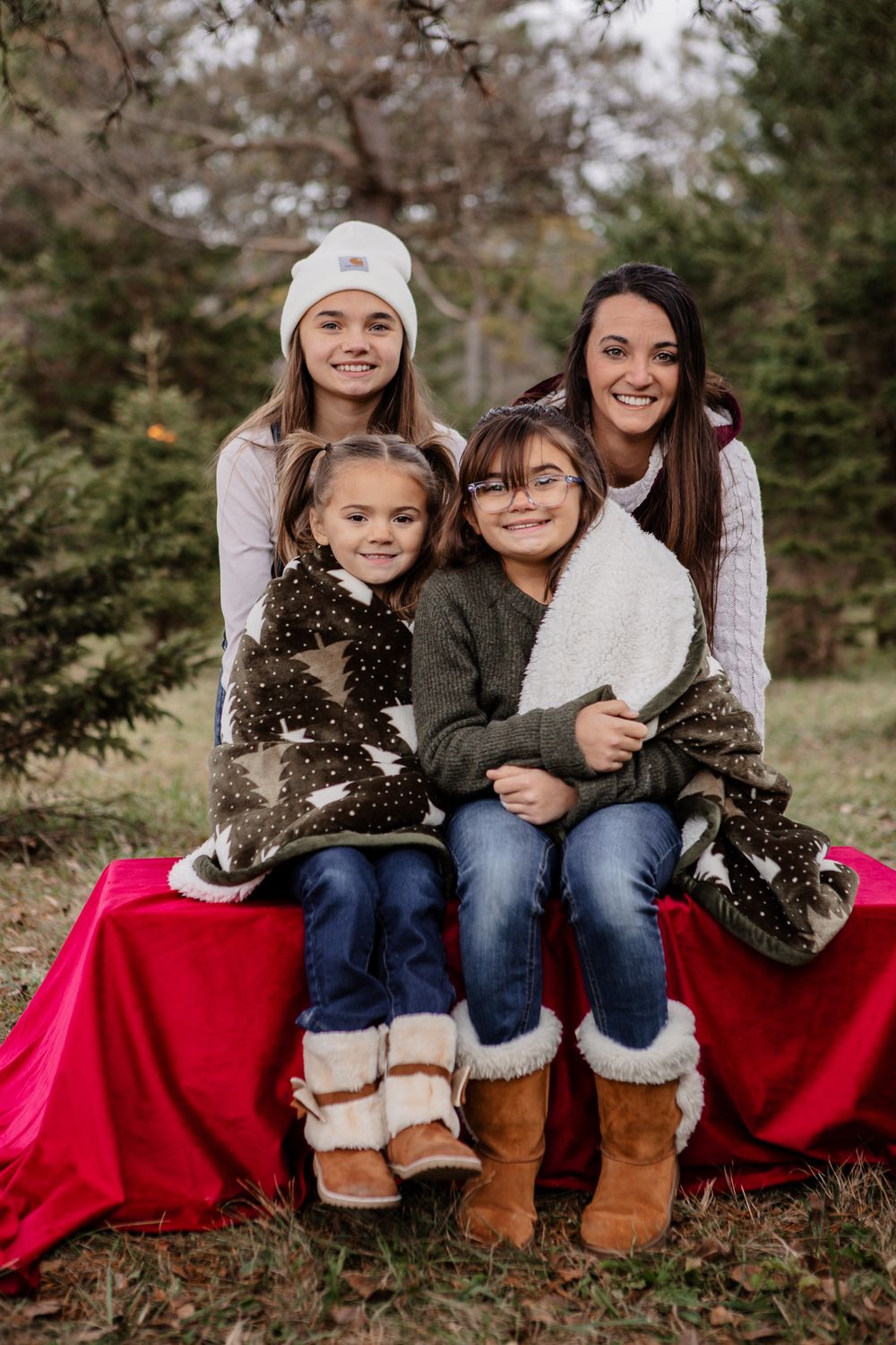 A cozy winter photo session with people sitting on a bright red blanket surrounded by evergreen trees.