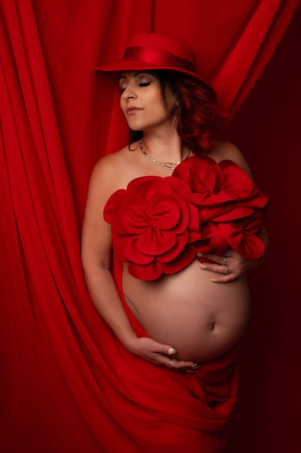 Artistic maternity photo with red hat and roses against dramatic red drapery background.