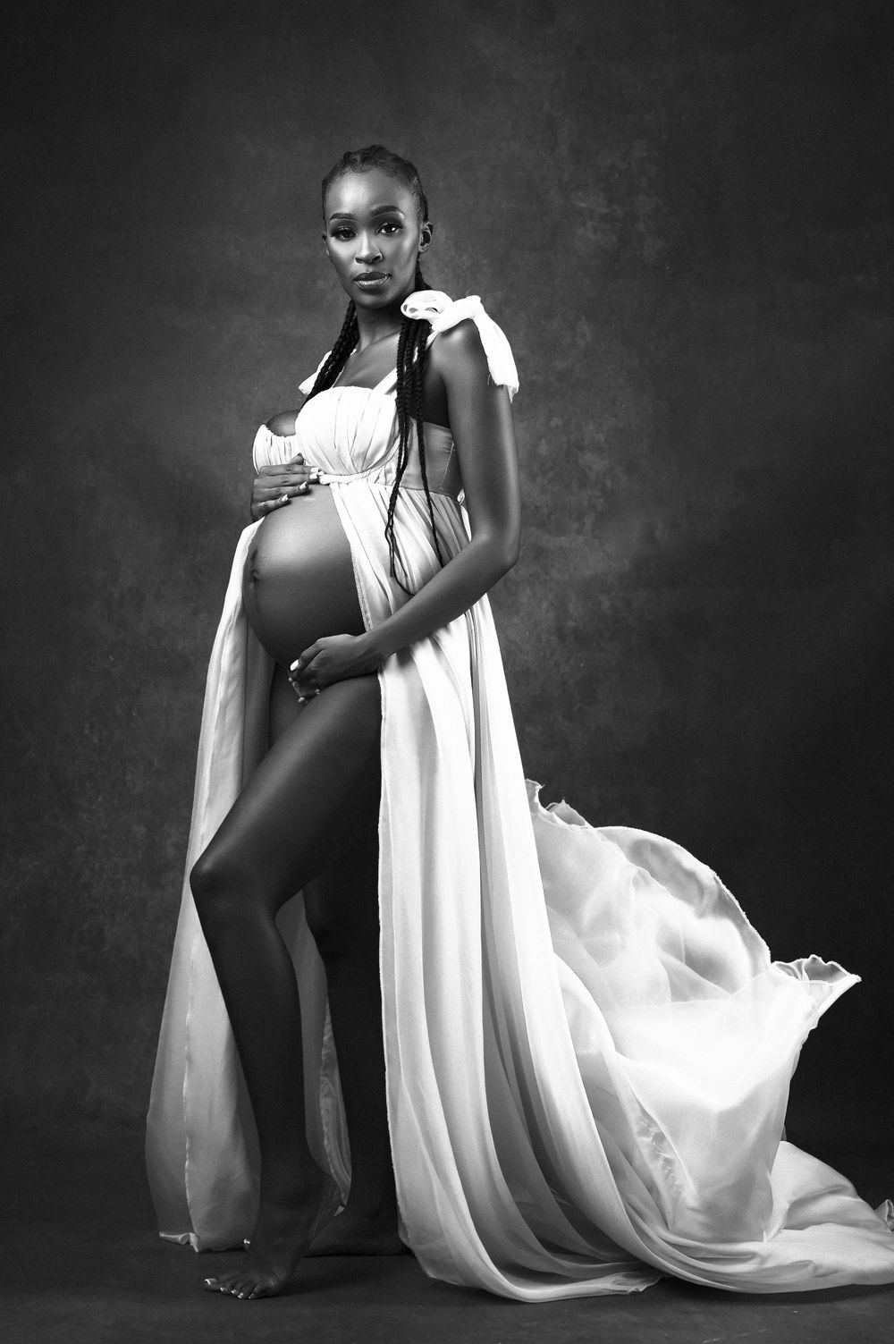 Artistic black and white maternity portrait featuring a flowing white dress against a dark backdrop.
