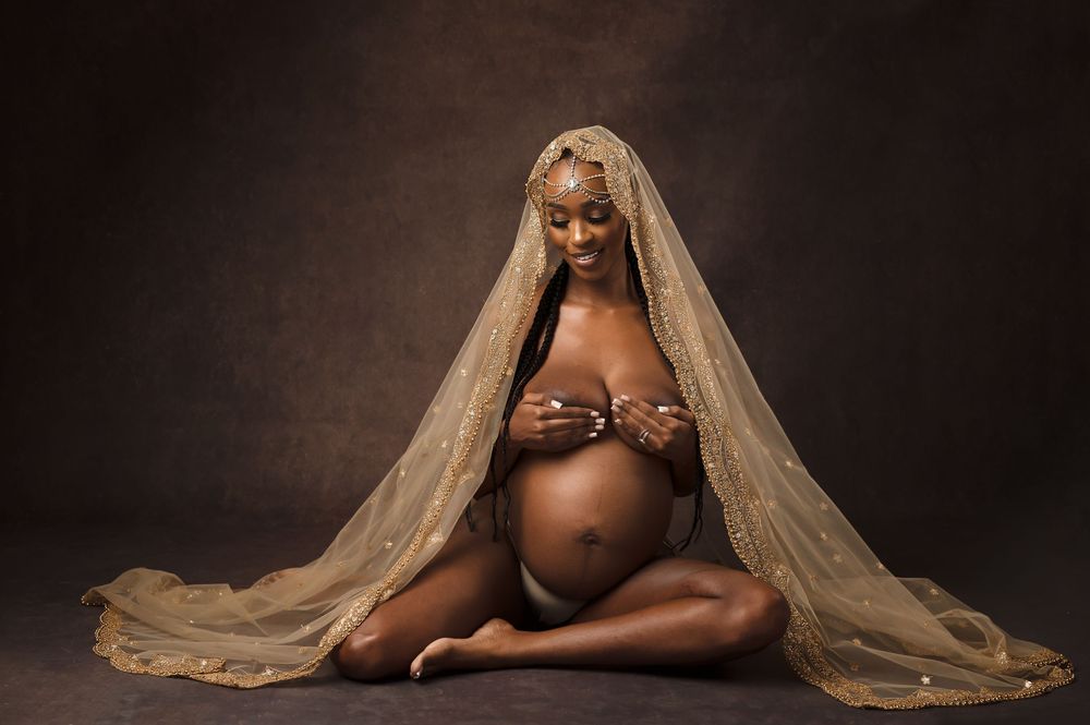 A pregnant person sits on the floor draped in flowing beige fabric against a dark studio backdrop.