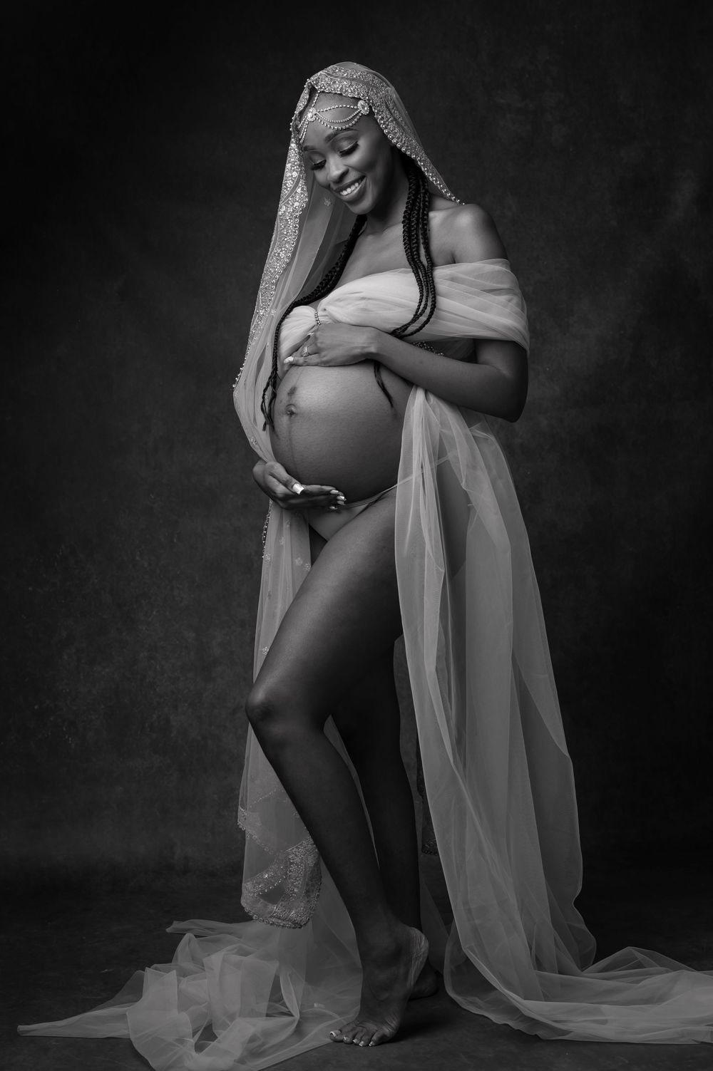 Artistic black and white maternity photo with flowing sheer fabric draping around an expectant mother in dramatic lighting.