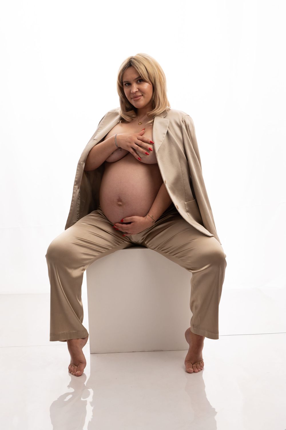 Artistic maternity photo showing a pregnant woman in beige clothing seated on white backdrop with dramatic lighting.