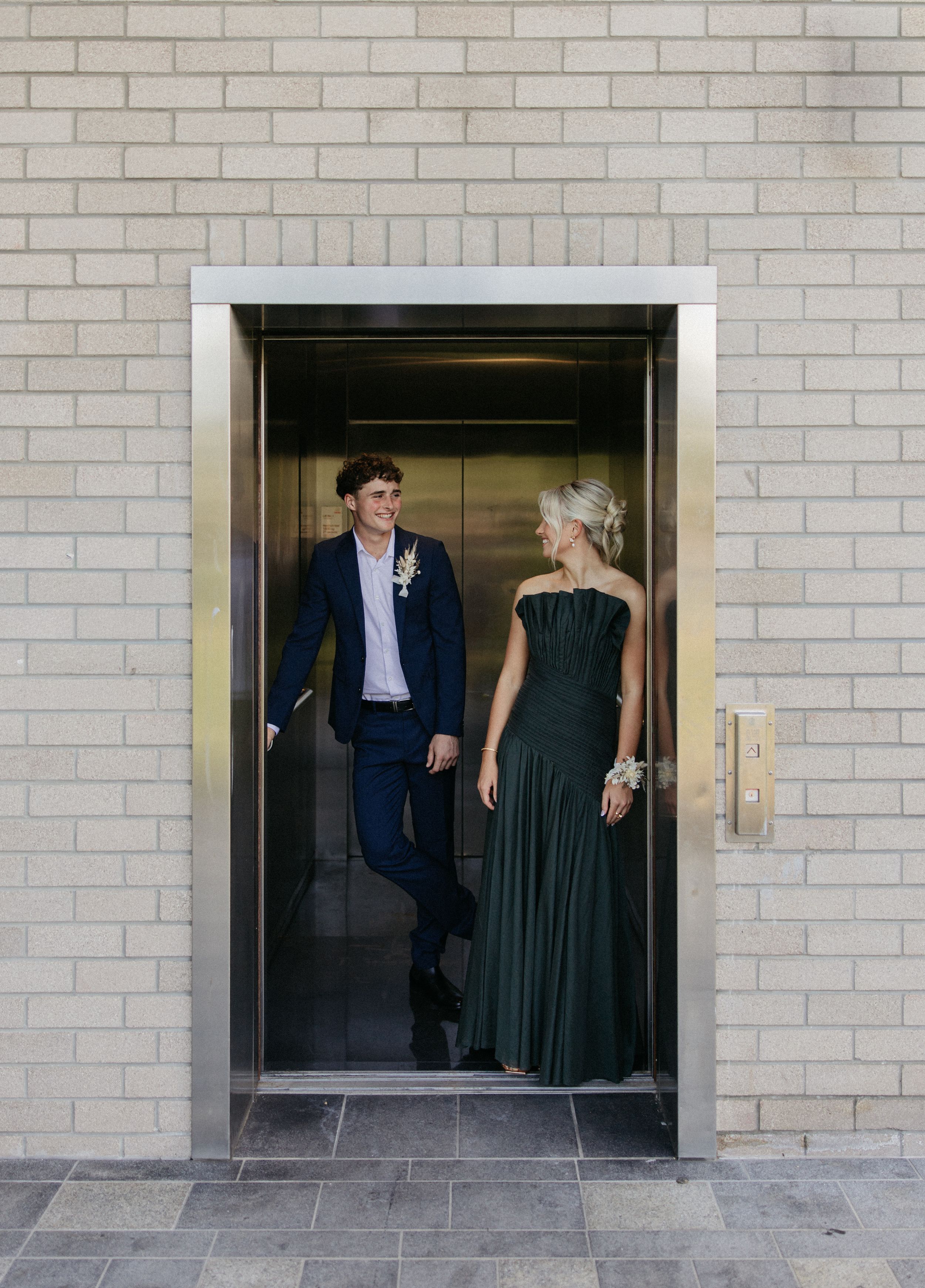 A couple in formal attire stands in an elevator, the man in a navy suit and woman in an emerald green gown, smiling at each other.