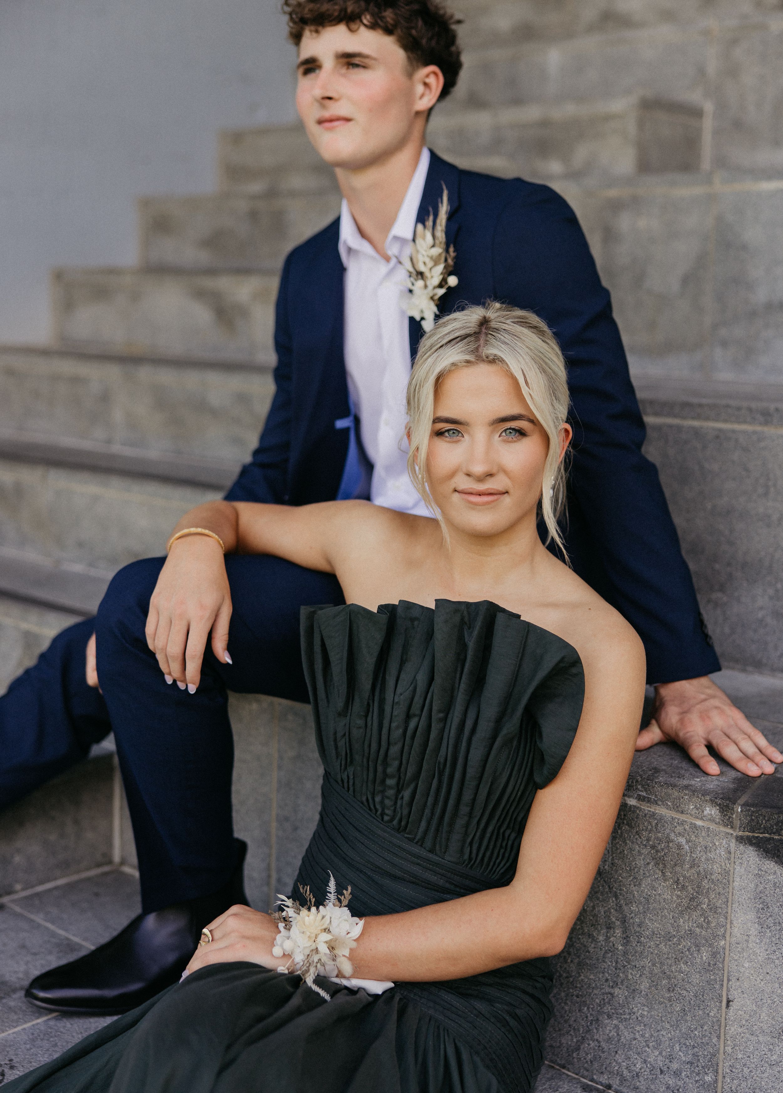 A young couple in formal prom attire pose on stone steps, wearing matching floral corsages.