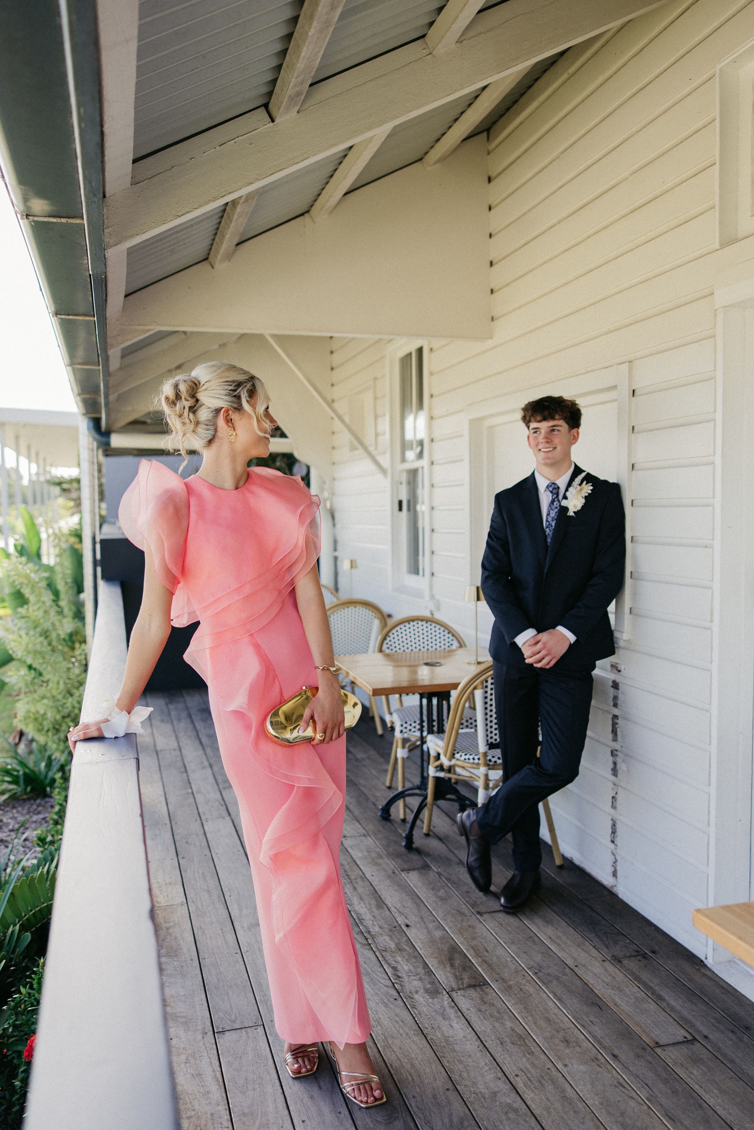 A woman in a pink gown and a man in a navy suit socializing on a wooden verandah of a white weatherboard building.
