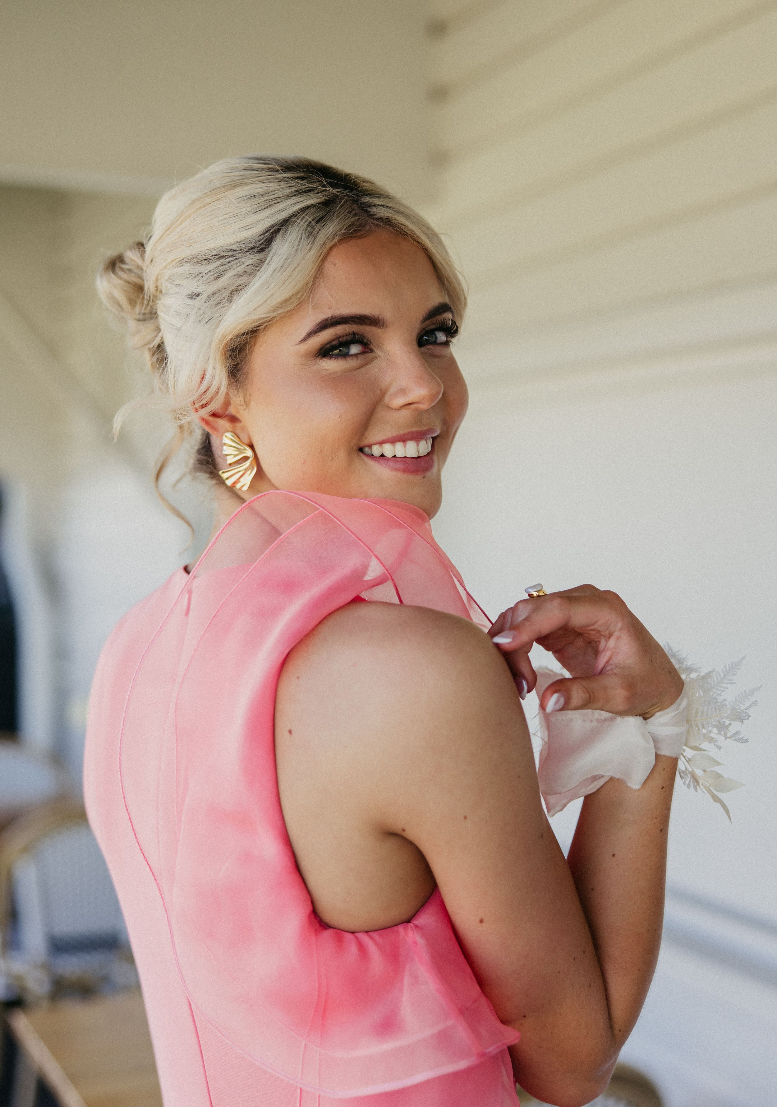 Smiling blonde woman in pink dress adjusting floral wrist corsage, wearing gold earrings.
