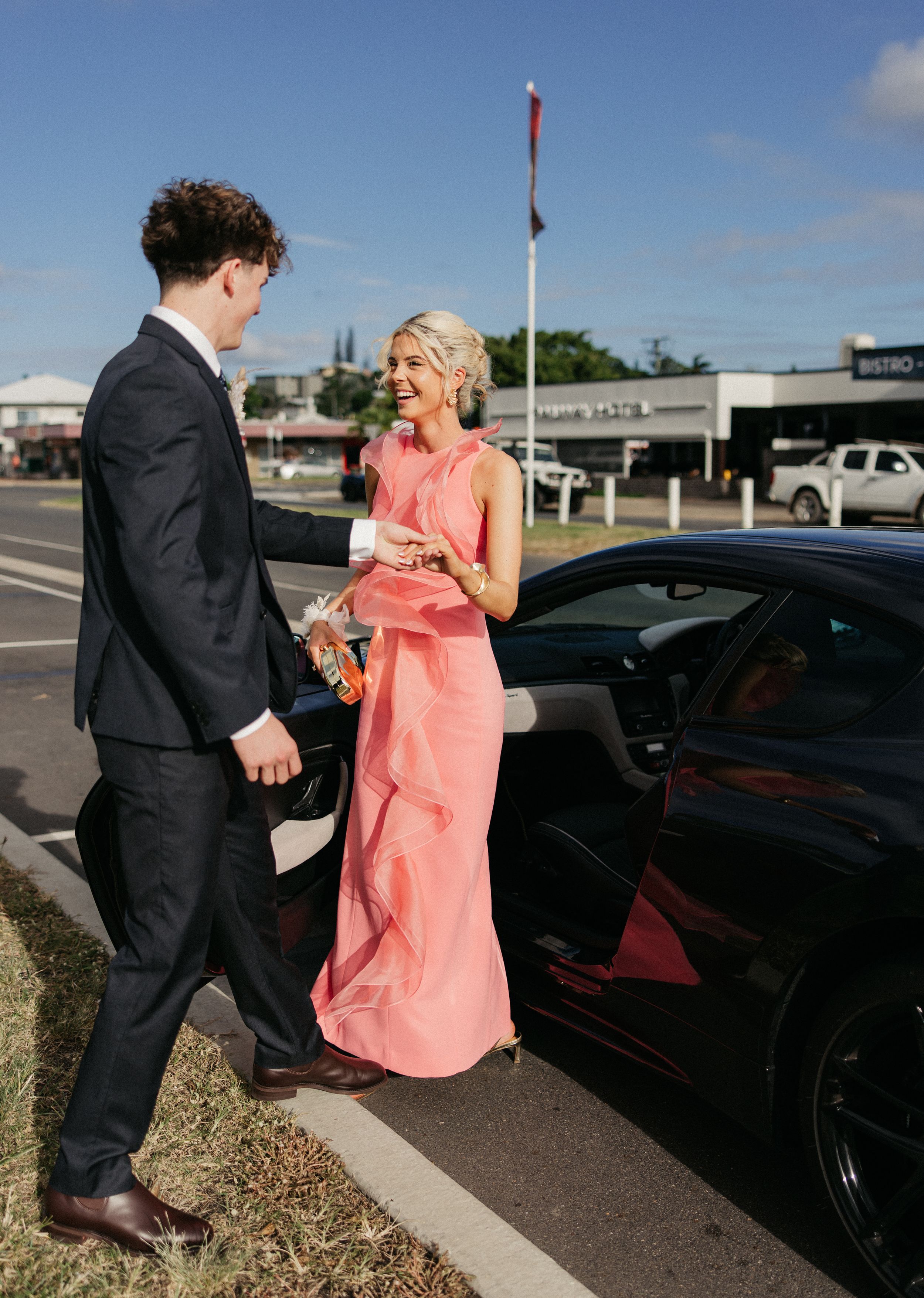 Couple in formal attire, man in dark suit, woman in pink gown, standing beside a black sports car outdoors.