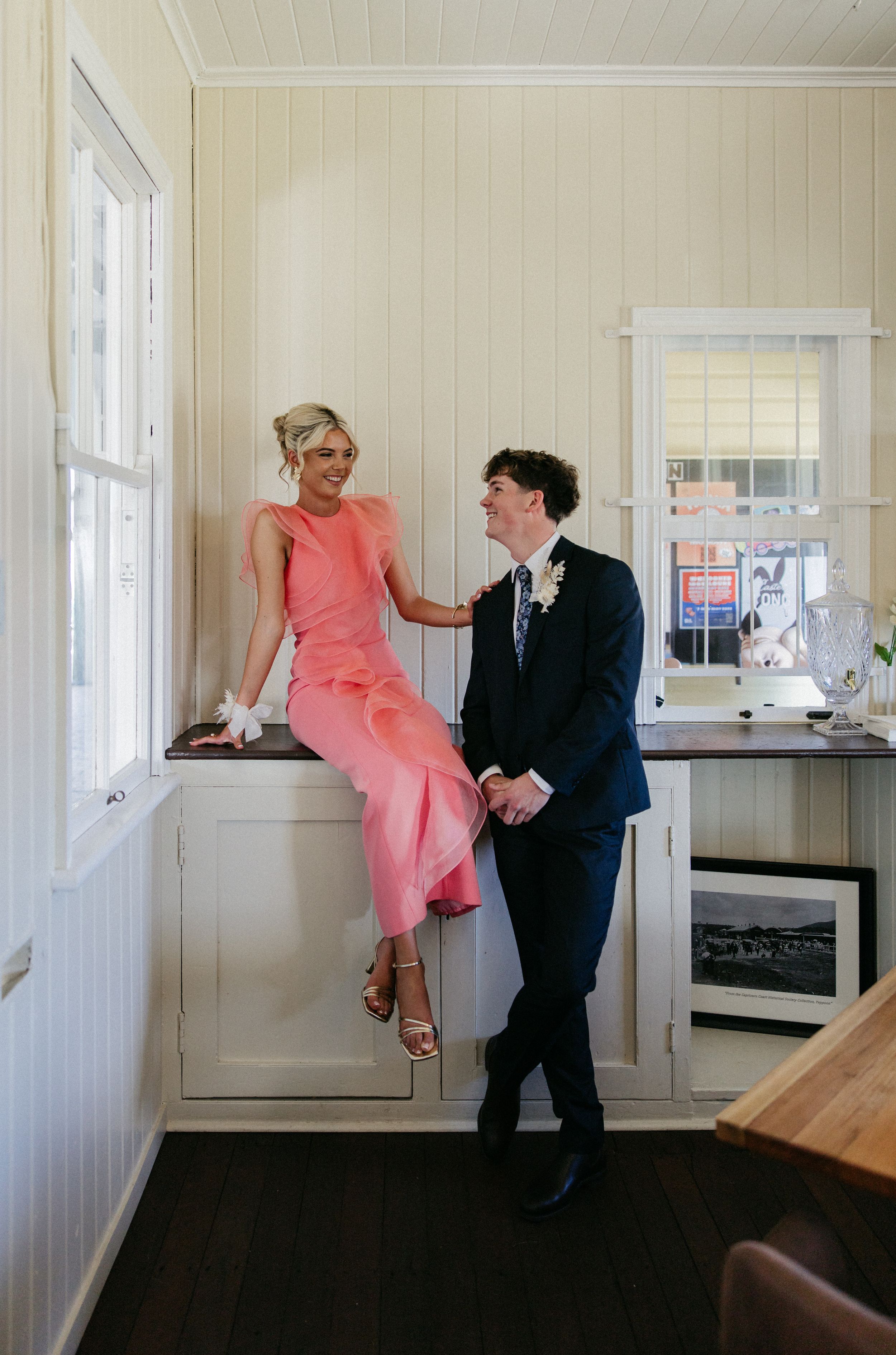 Couple in formal attire laughing together, woman in coral pink dress seated on counter, man in navy suit beside her.