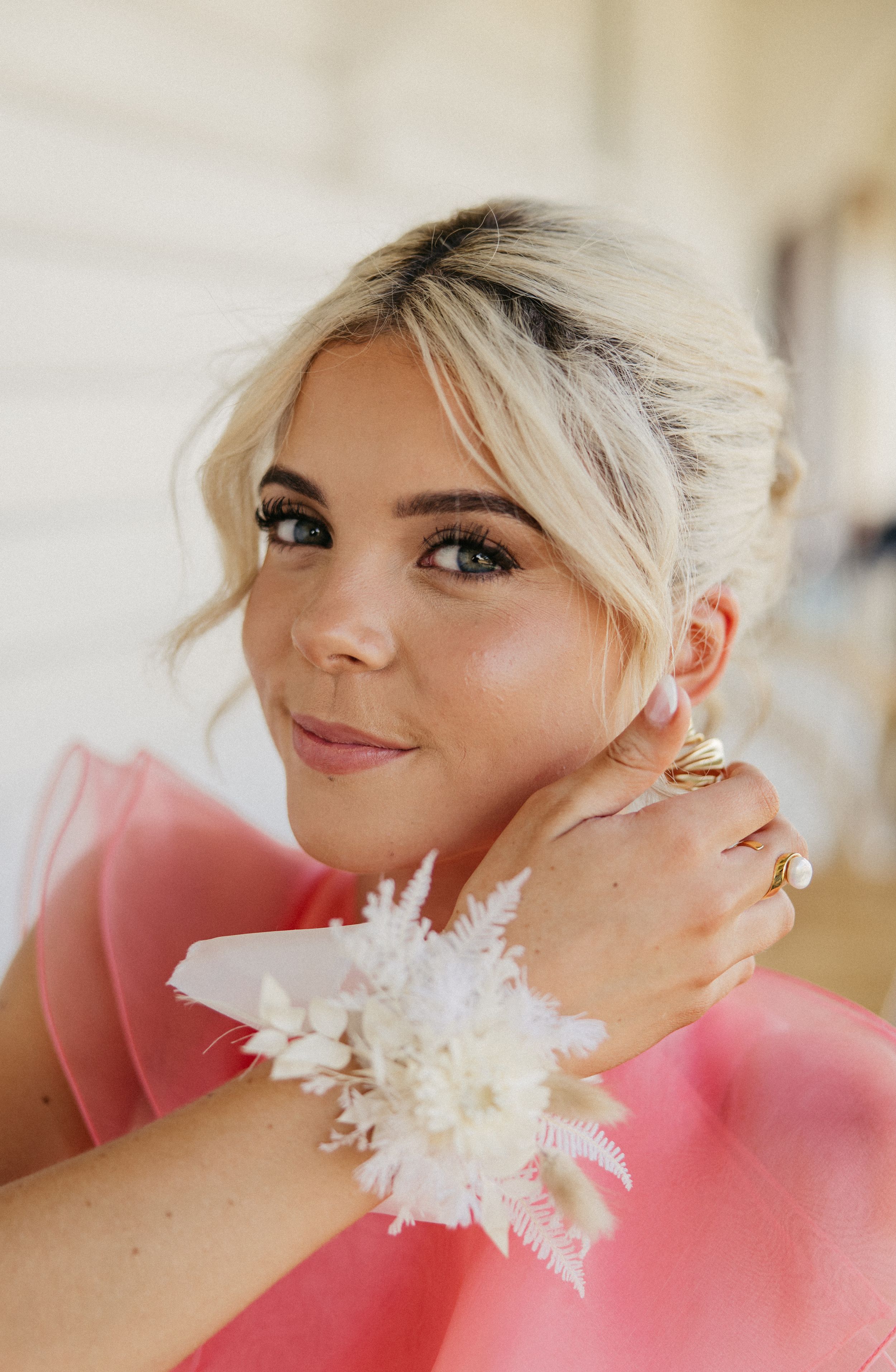 Blonde woman in pink dress wearing a white floral wrist corsage, smiling softly with an elegant updo hairstyle.