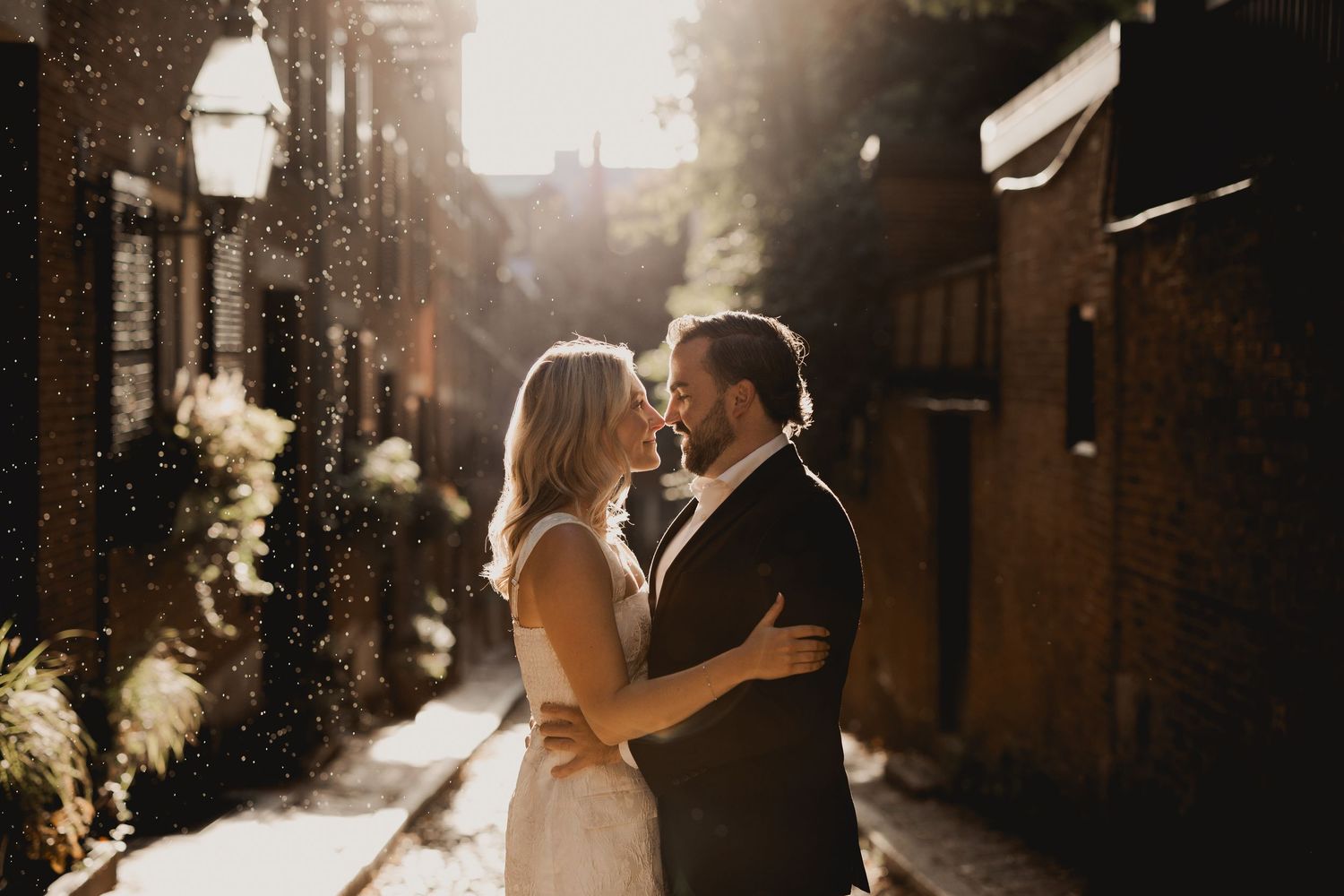 A couple embraces on a snowy street at sunset with brick buildings in the background.