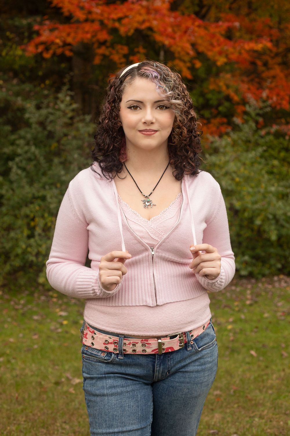 A person wearing a pink sweater and jeans poses outdoors against autumn foliage in a casual photoshoot series.