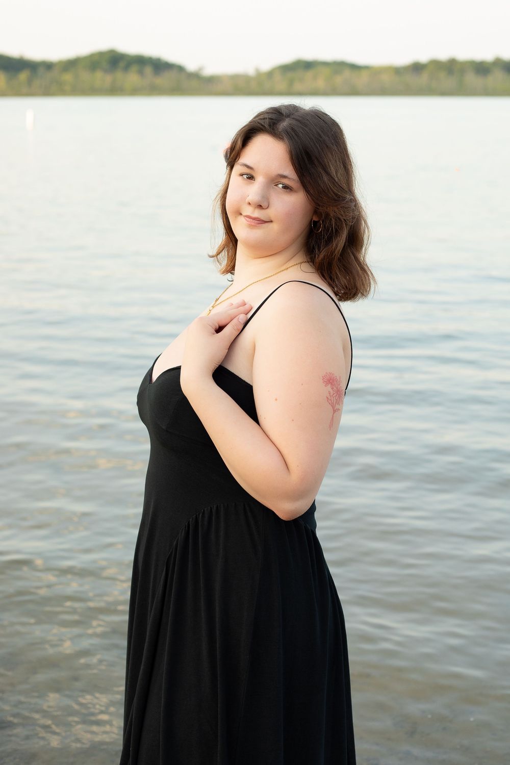 Woman in black dress smiling over shoulder during maternity photoshoot by waterfront.