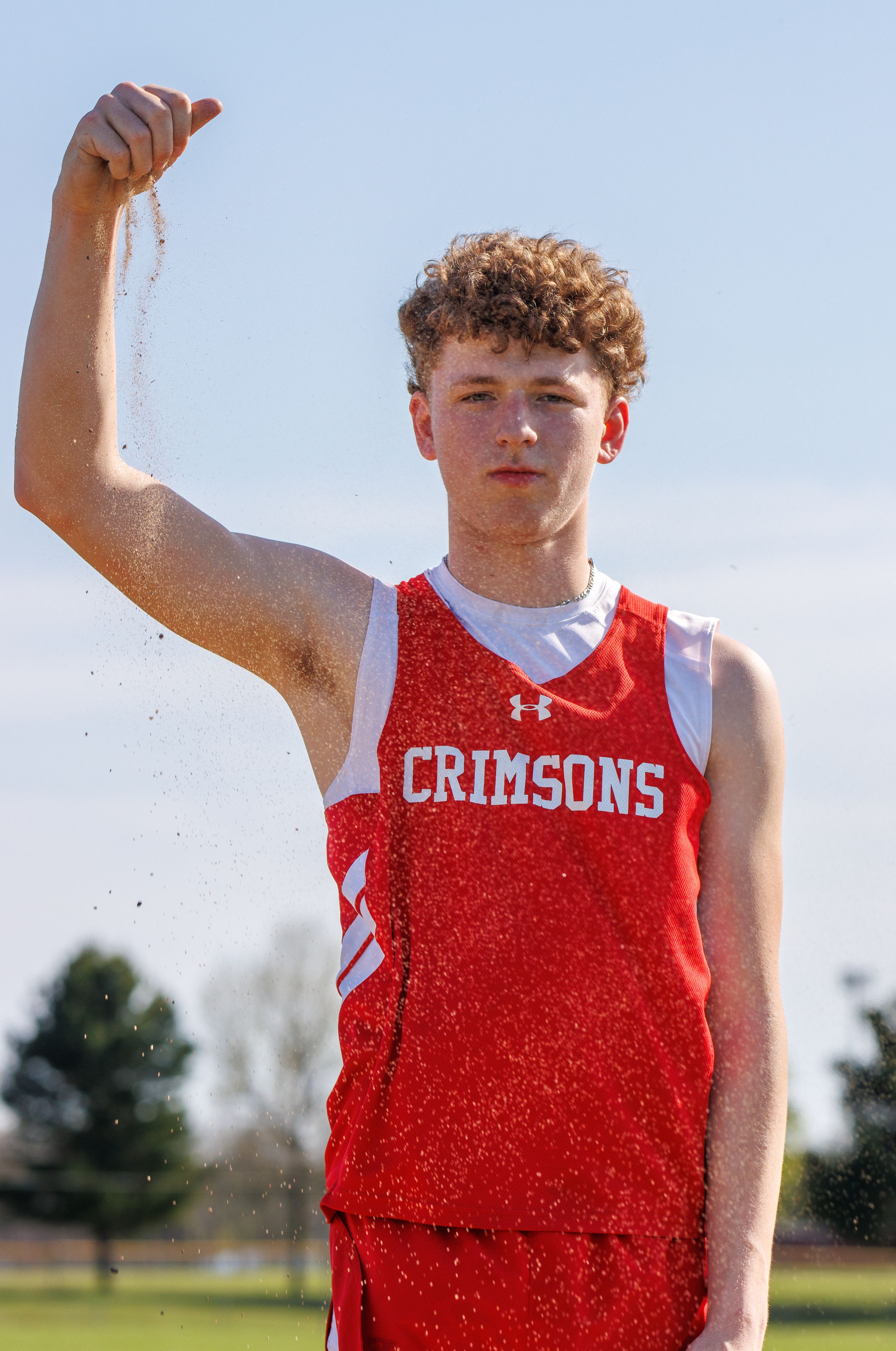 An athlete in a red Crimsons jersey holds up sand against a blue sky.