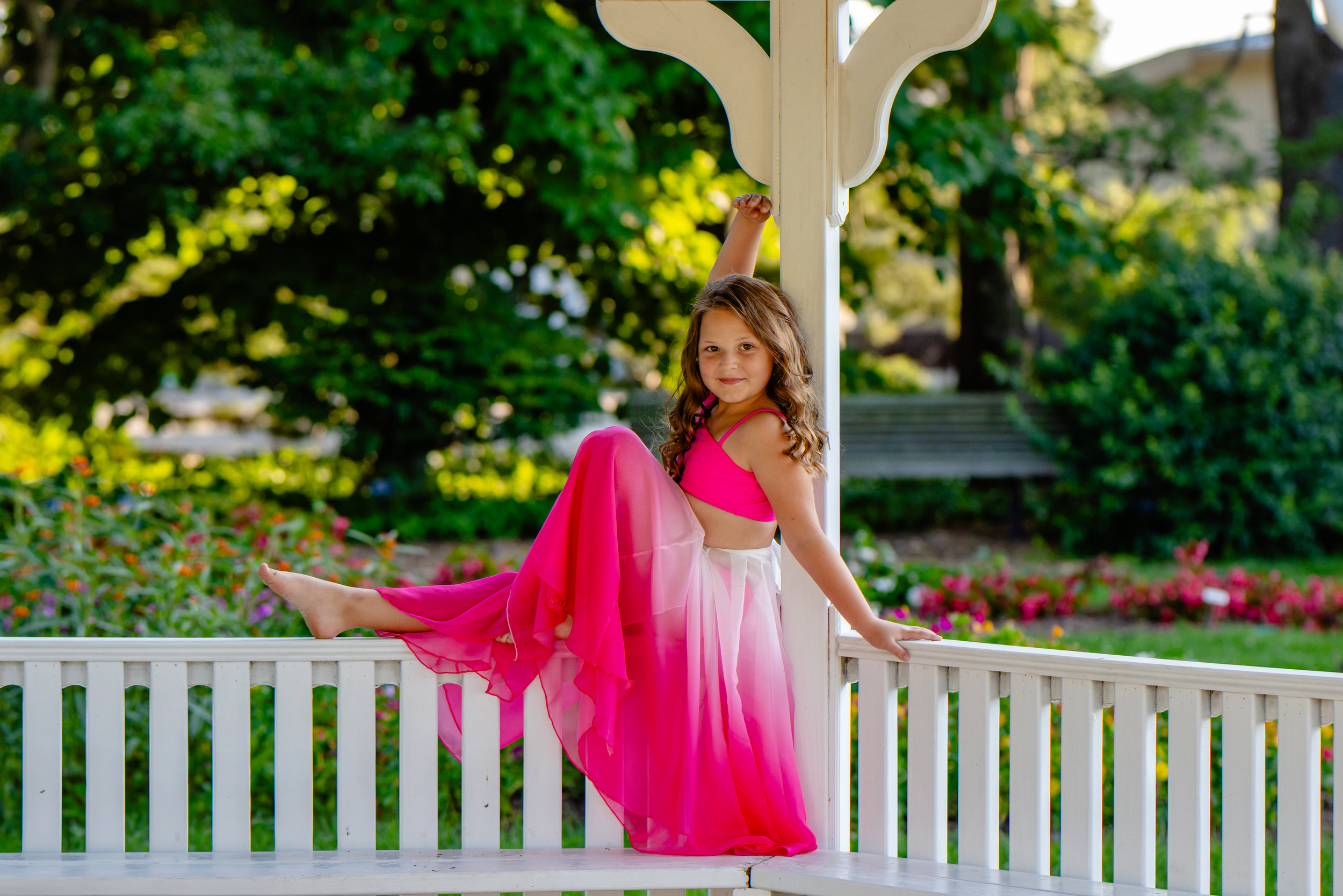 A girl in a flowing pink dress twirls on a white gazebo in a garden setting.