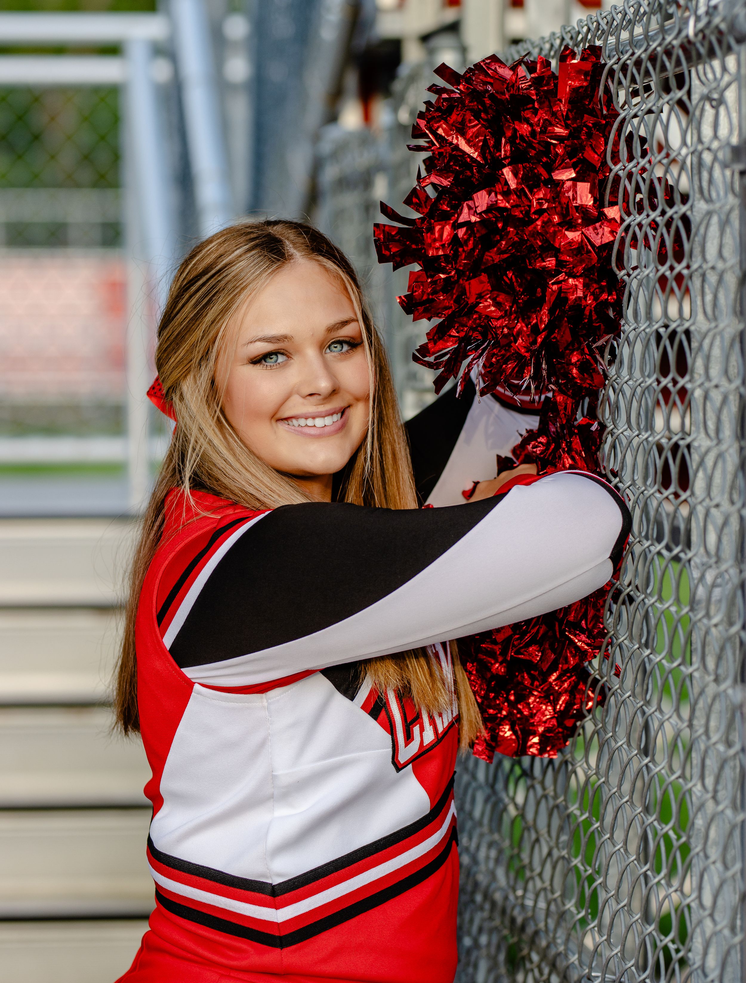 A cheerleader in red and white uniform poses with pom poms against chain link fence.