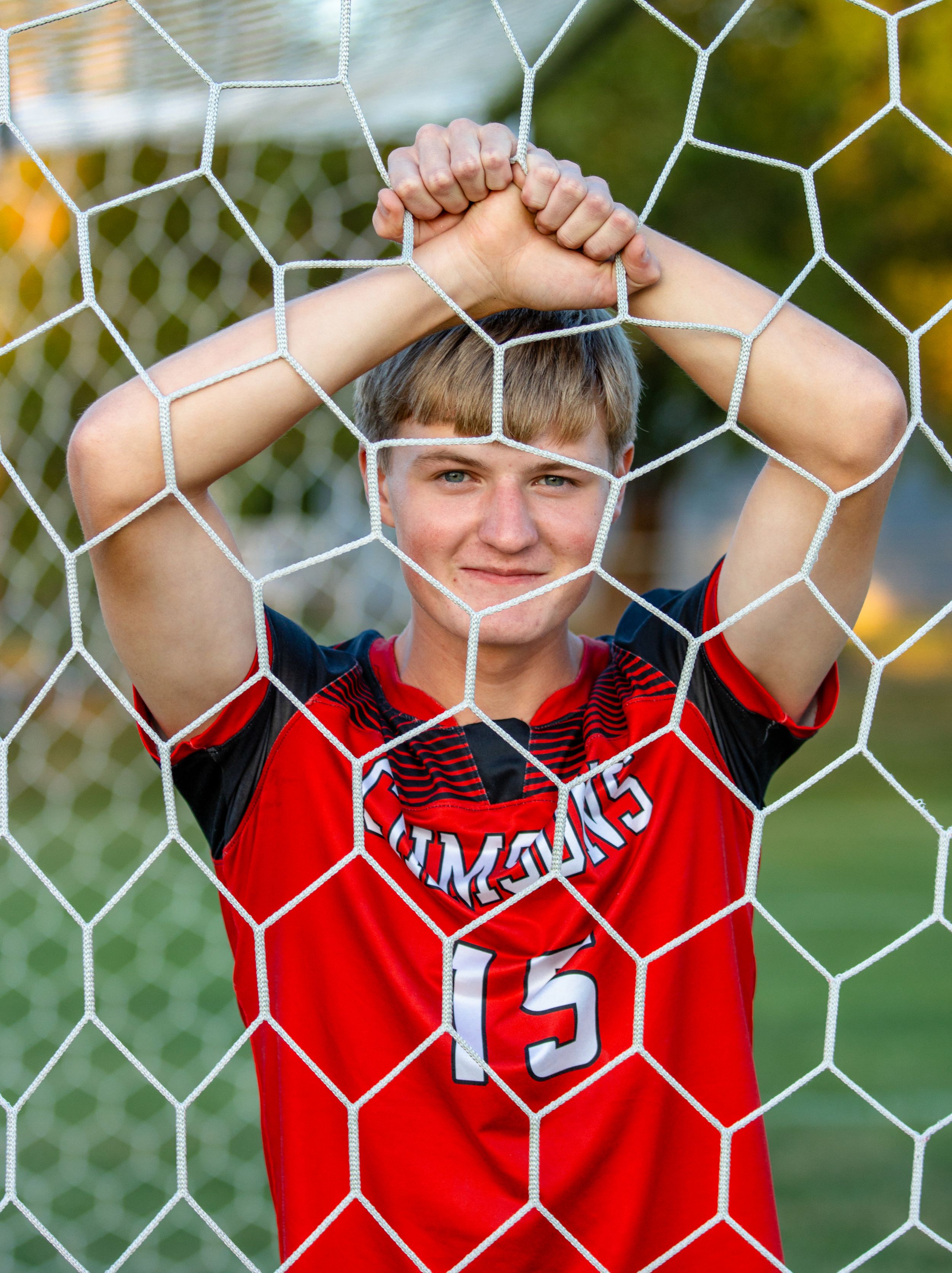 A youth athlete in red jersey poses behind a soccer goal net.