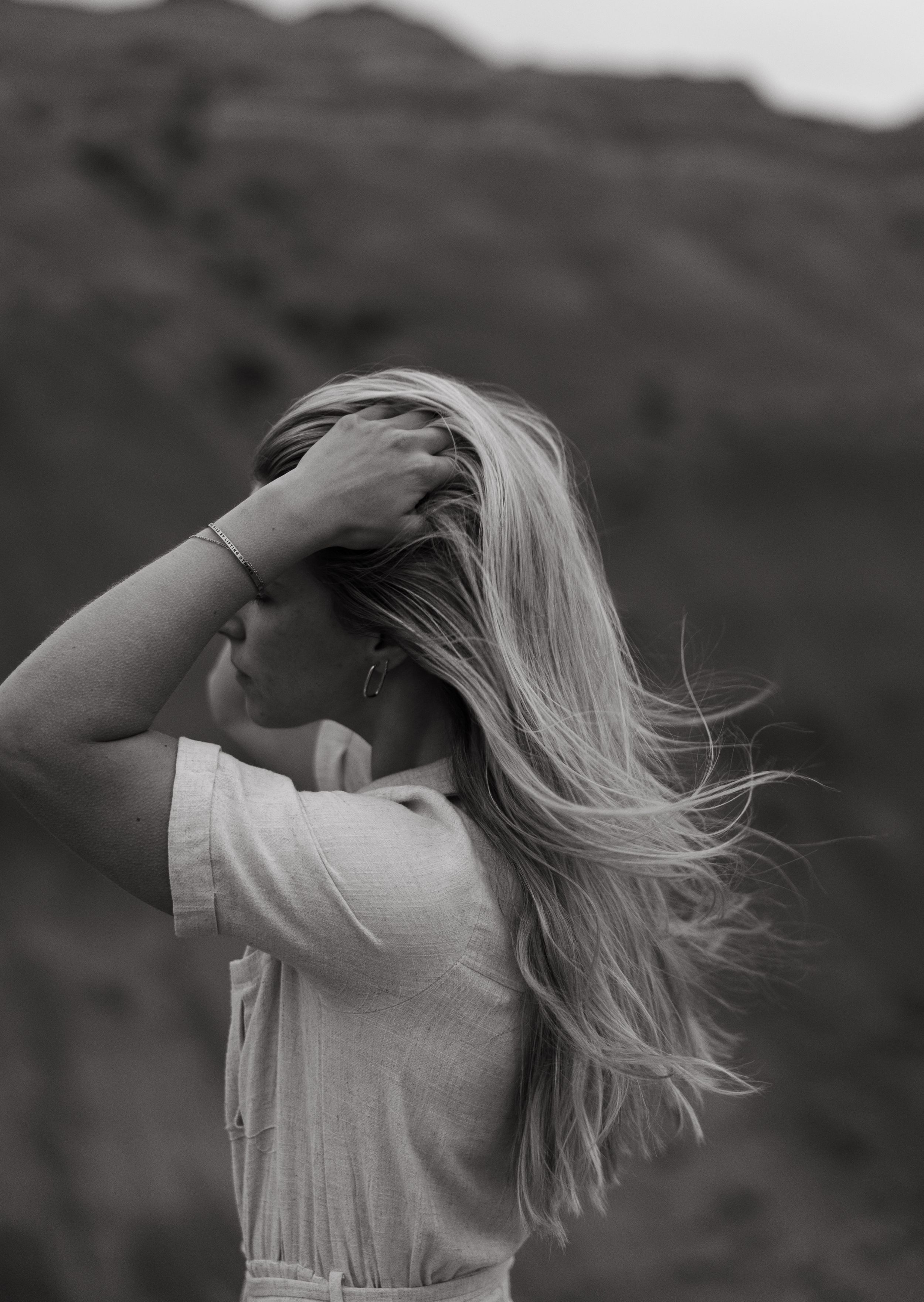 Black and white portrait of blonde woman with windswept hair against mountain backdrop.