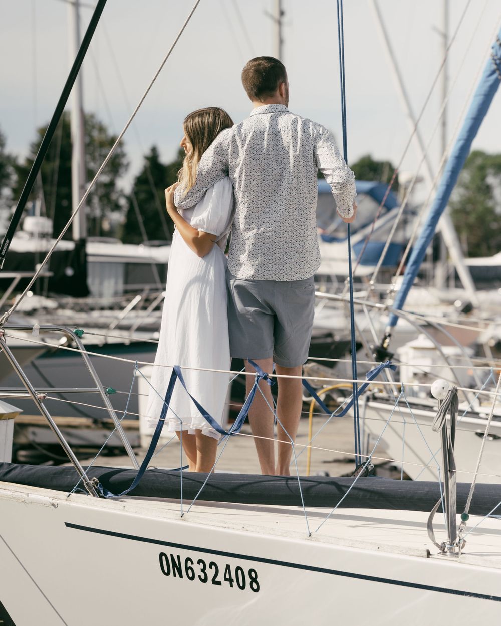 A couple stands together on a white sailboat deck at a marina on a sunny day.