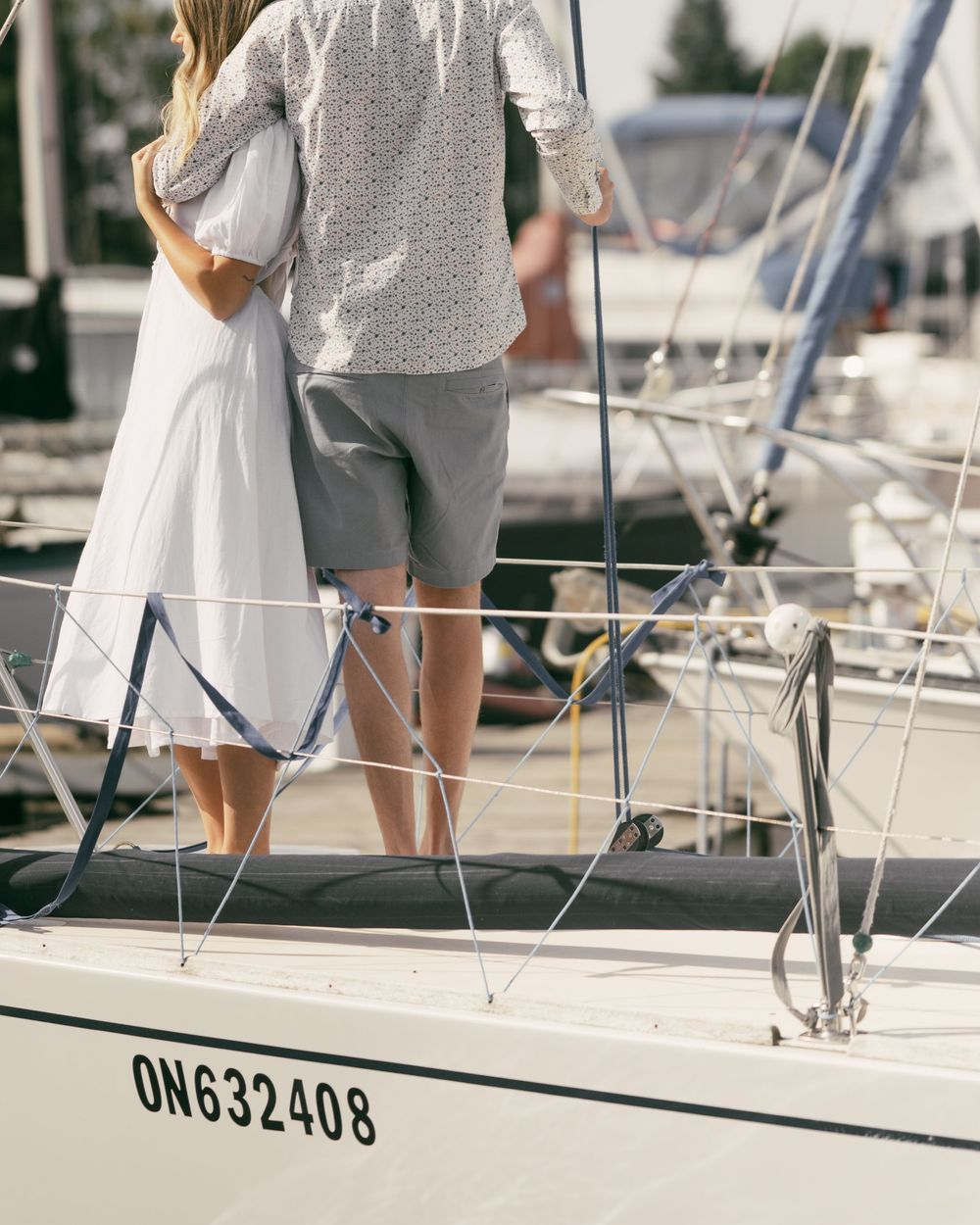 A couple in casual summer attire stands on a white sailboat with marina views.