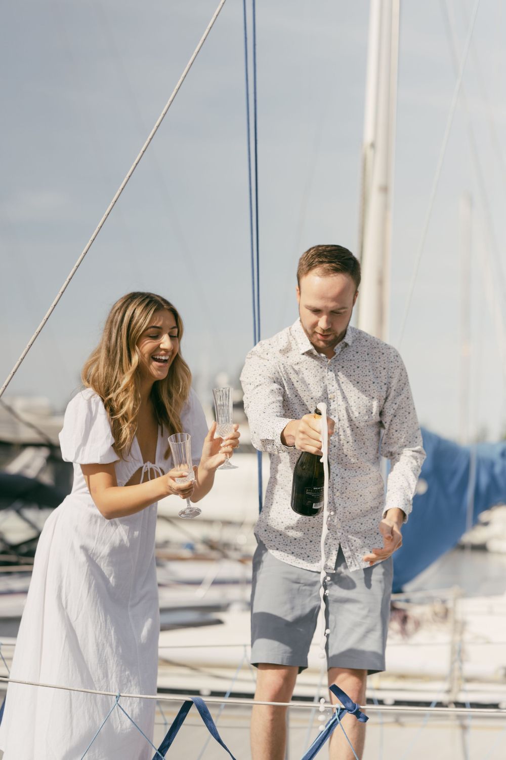 A couple shares champagne and laughter on a sailboat deck in a marina.