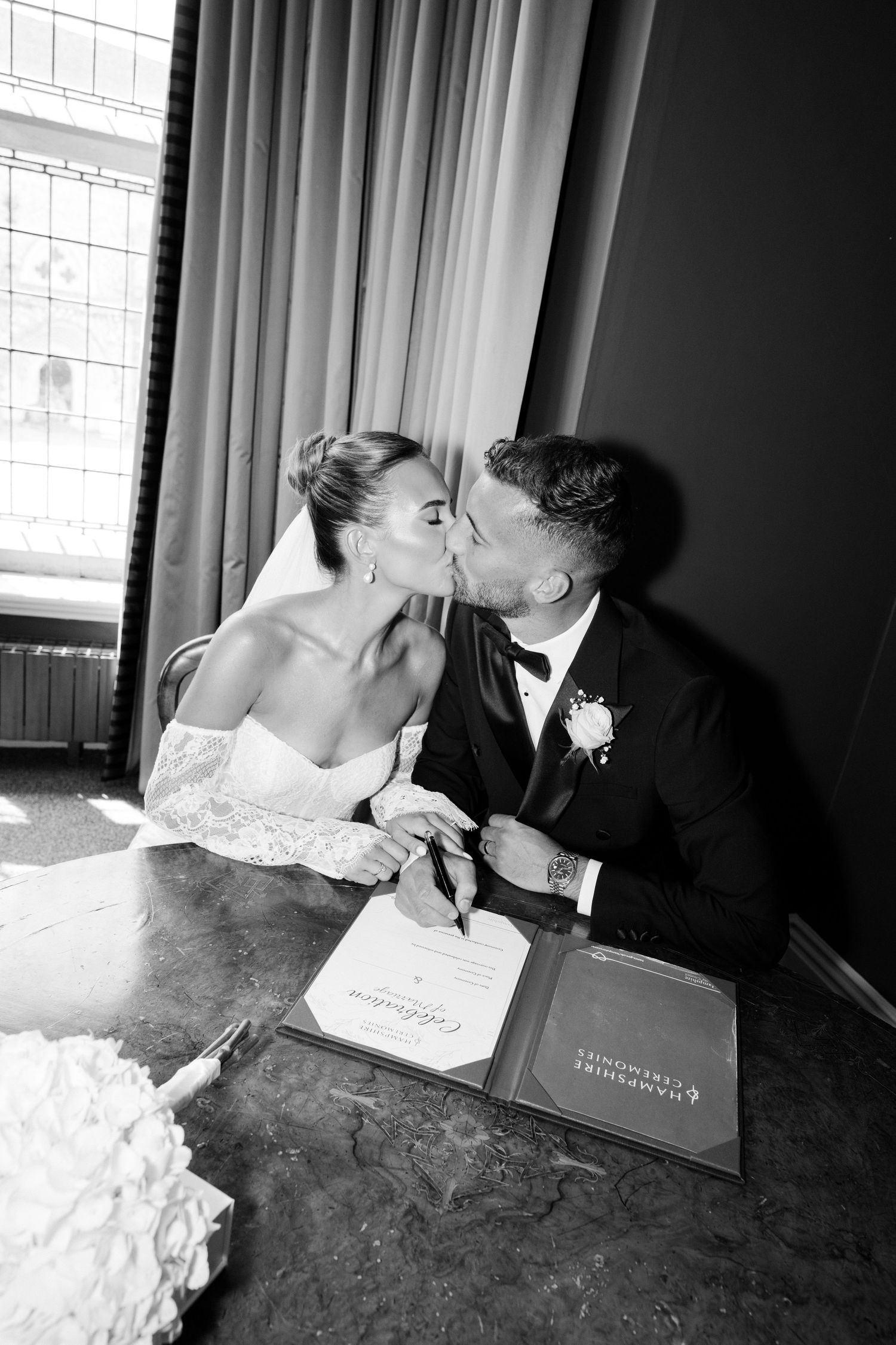 A black and white wedding photo showing a couple sharing a kiss while signing their marriage documents.