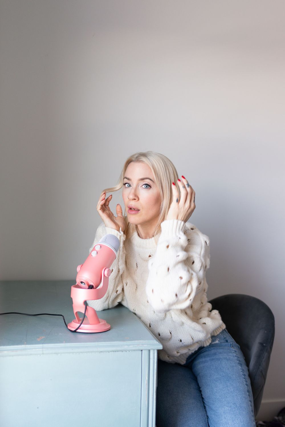 Person in white sweater sitting at desk with pink microphone, recording podcast or audio content while listening.