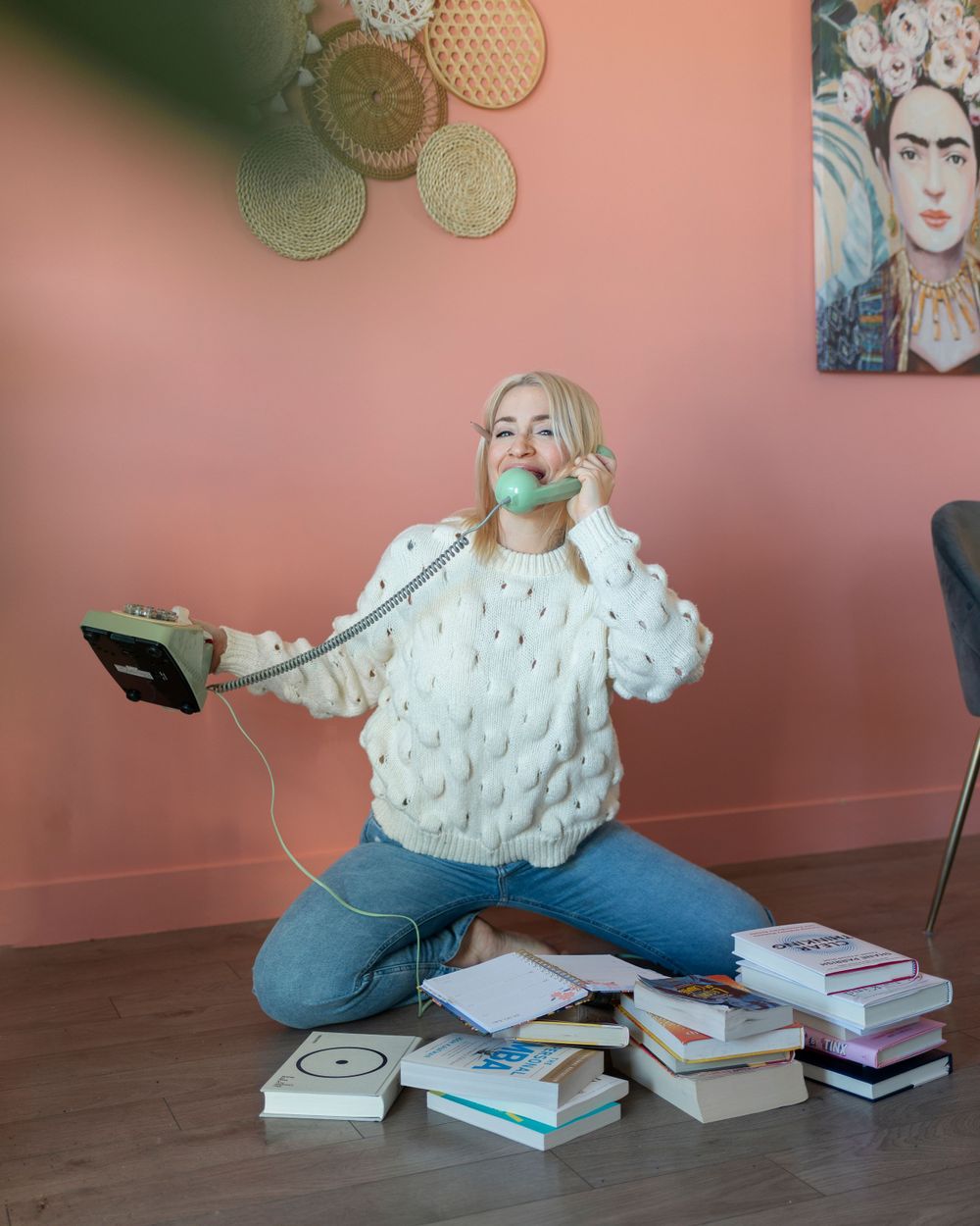 Person in white sweater sits cross-legged on floor with stacks of books and device with headphones around neck.