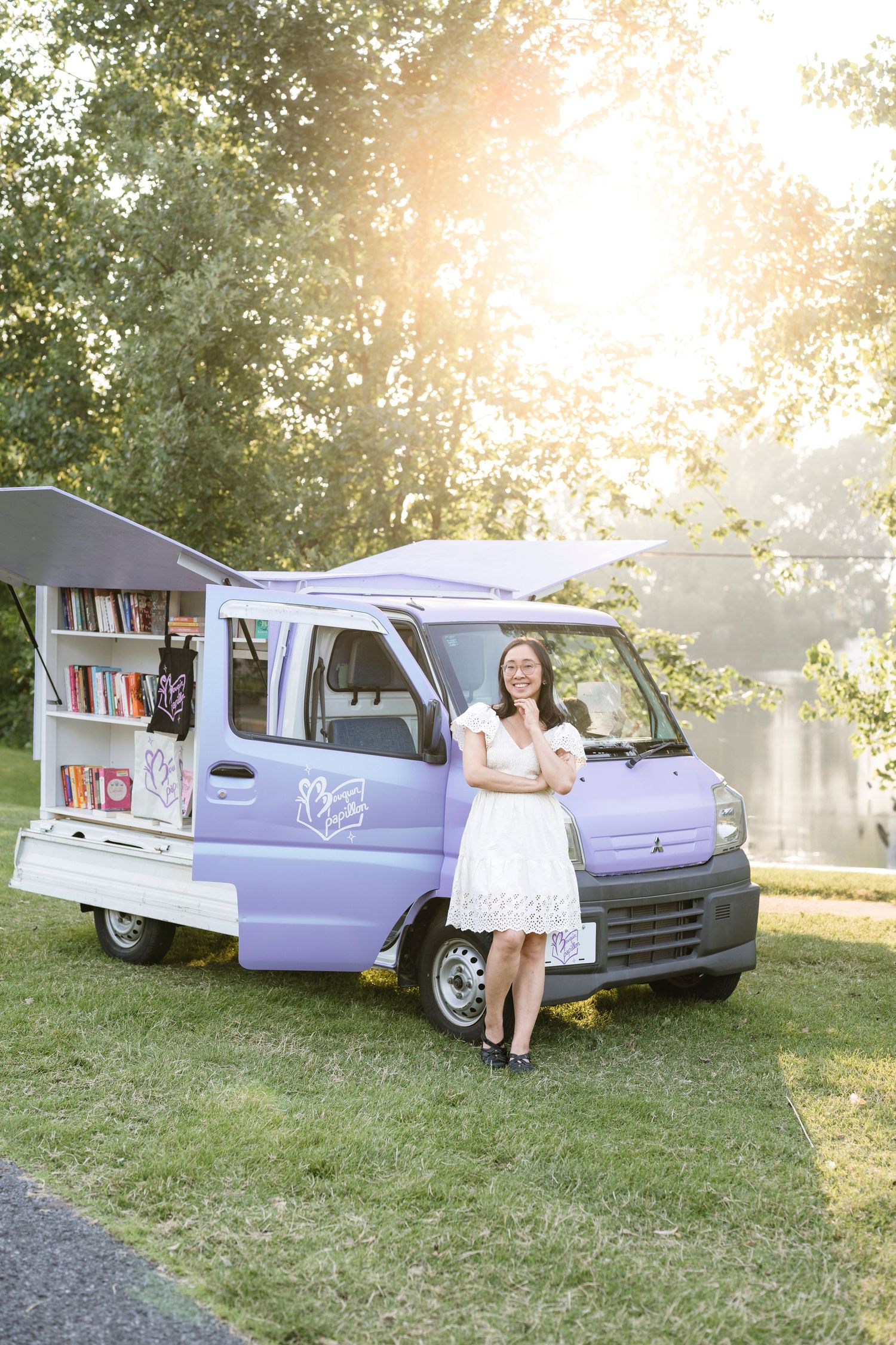 A mobile library van painted in light purple sits on grass during golden hour with sunlight filtering through trees.