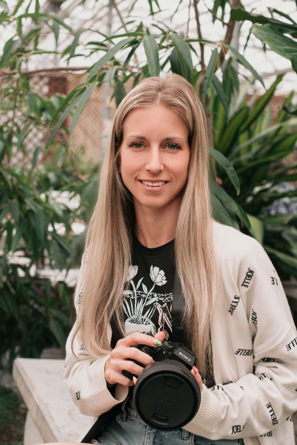 A photographer in a white sweater holds a professional camera while posing against a leafy green backdrop.