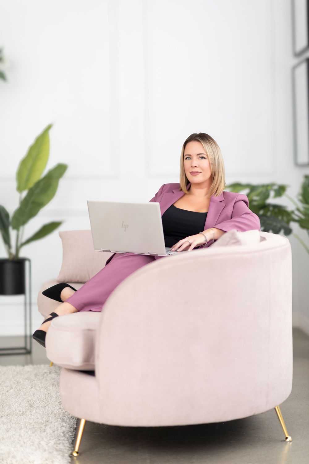 Woman in pink sweater works on laptop while relaxing on curved blush pink modern accent chair in bright room.