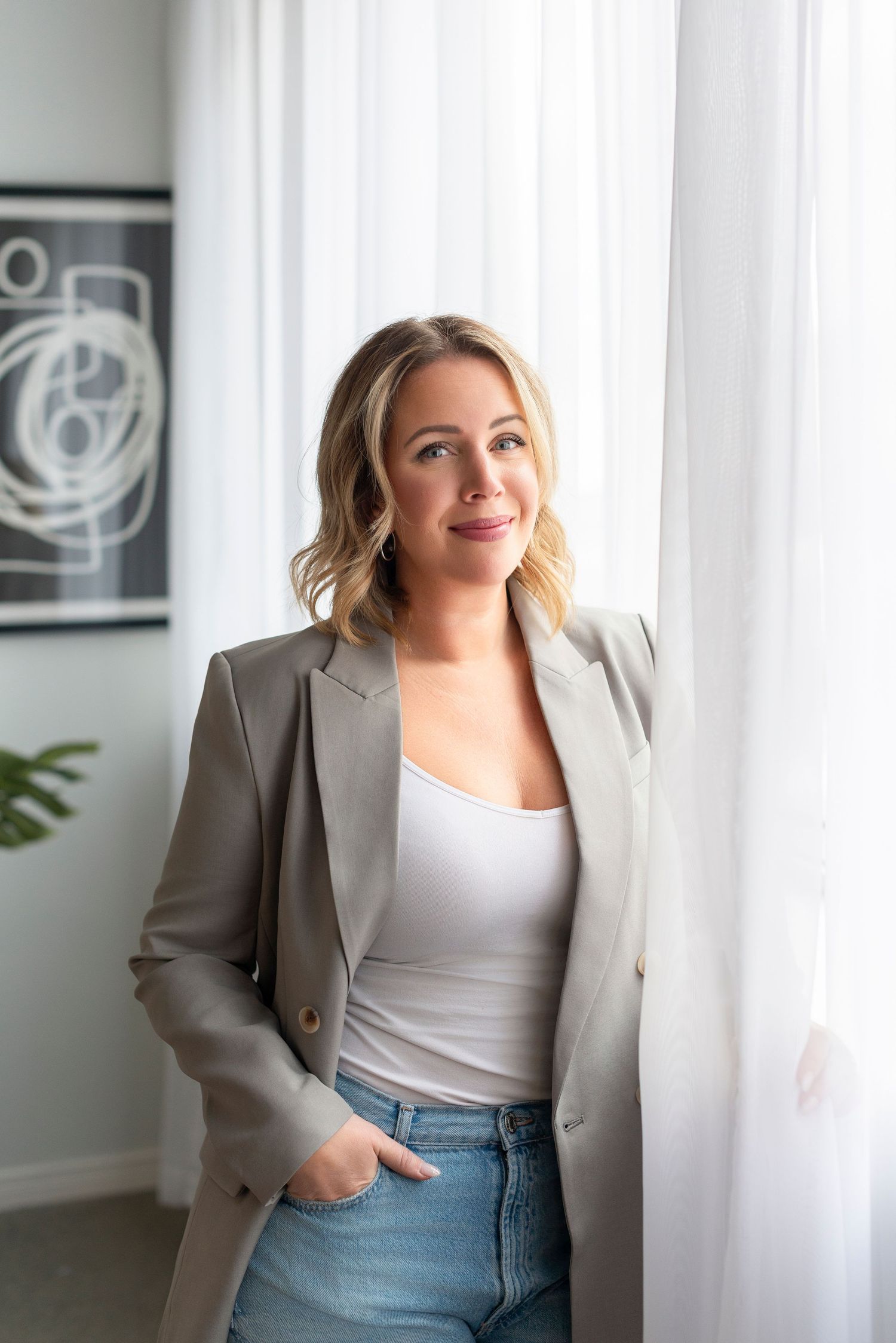 Professional woman in beige blazer and white top poses against white background in modern office setting.