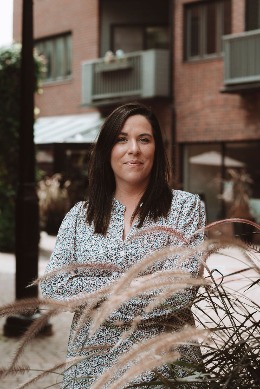 Professional portrait featuring a floral patterned dress against a brick building with decorative grasses in foreground.
