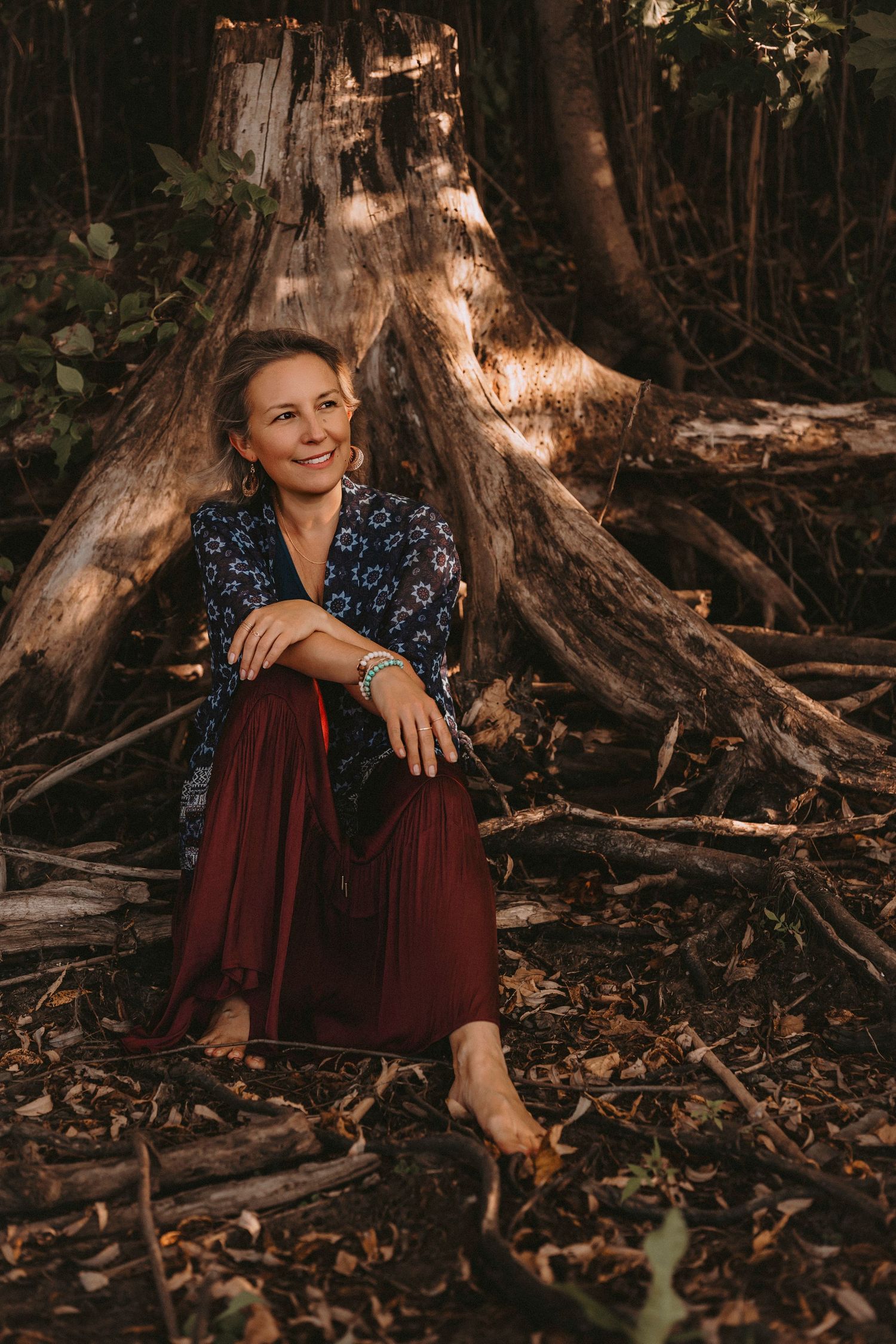 A person in a floral navy top and burgundy skirt poses in a dreamy forest setting with sunlit trees.