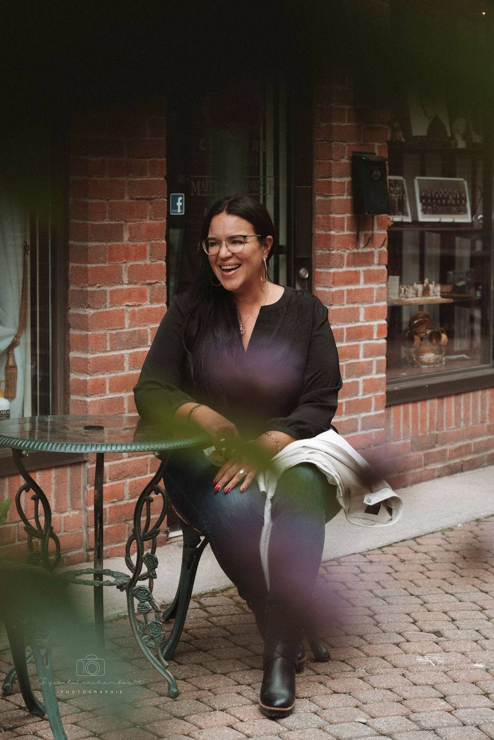 Casual outdoor portrait at a brick storefront with woman seated at bistro table in black attire.