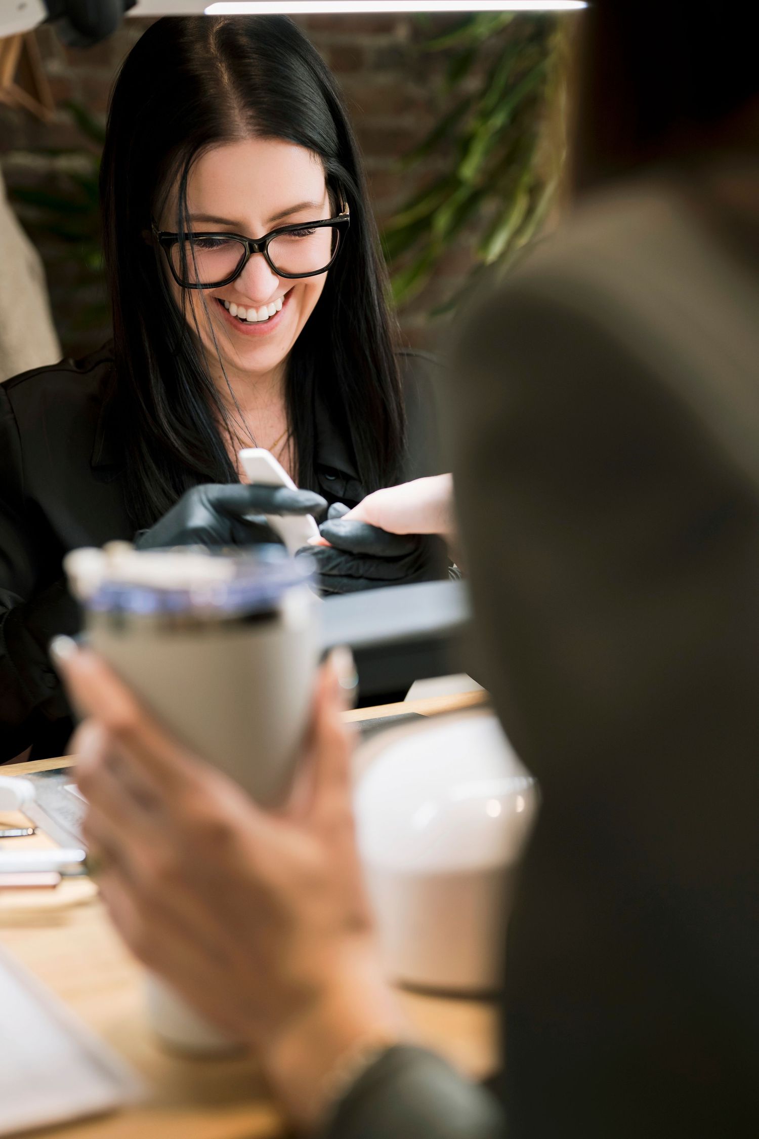 A person with long dark hair and glasses smiles while working at a desk with papers and a coffee cup.