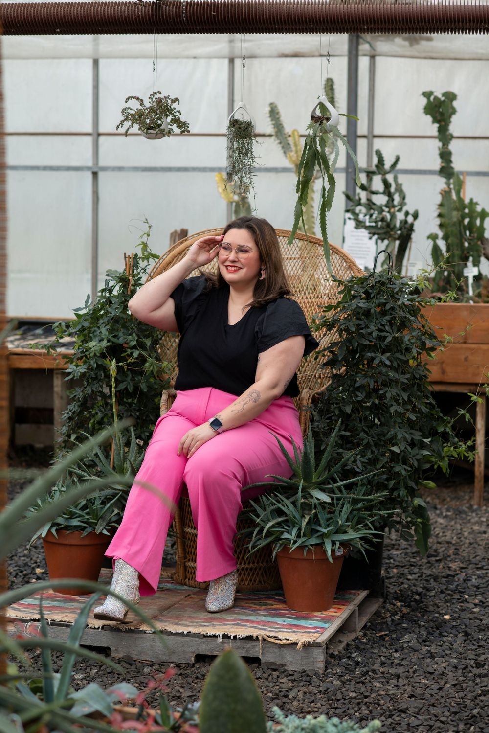 A person in a black top and bright pink pants sits on a chair surrounded by plants in a greenhouse setting.