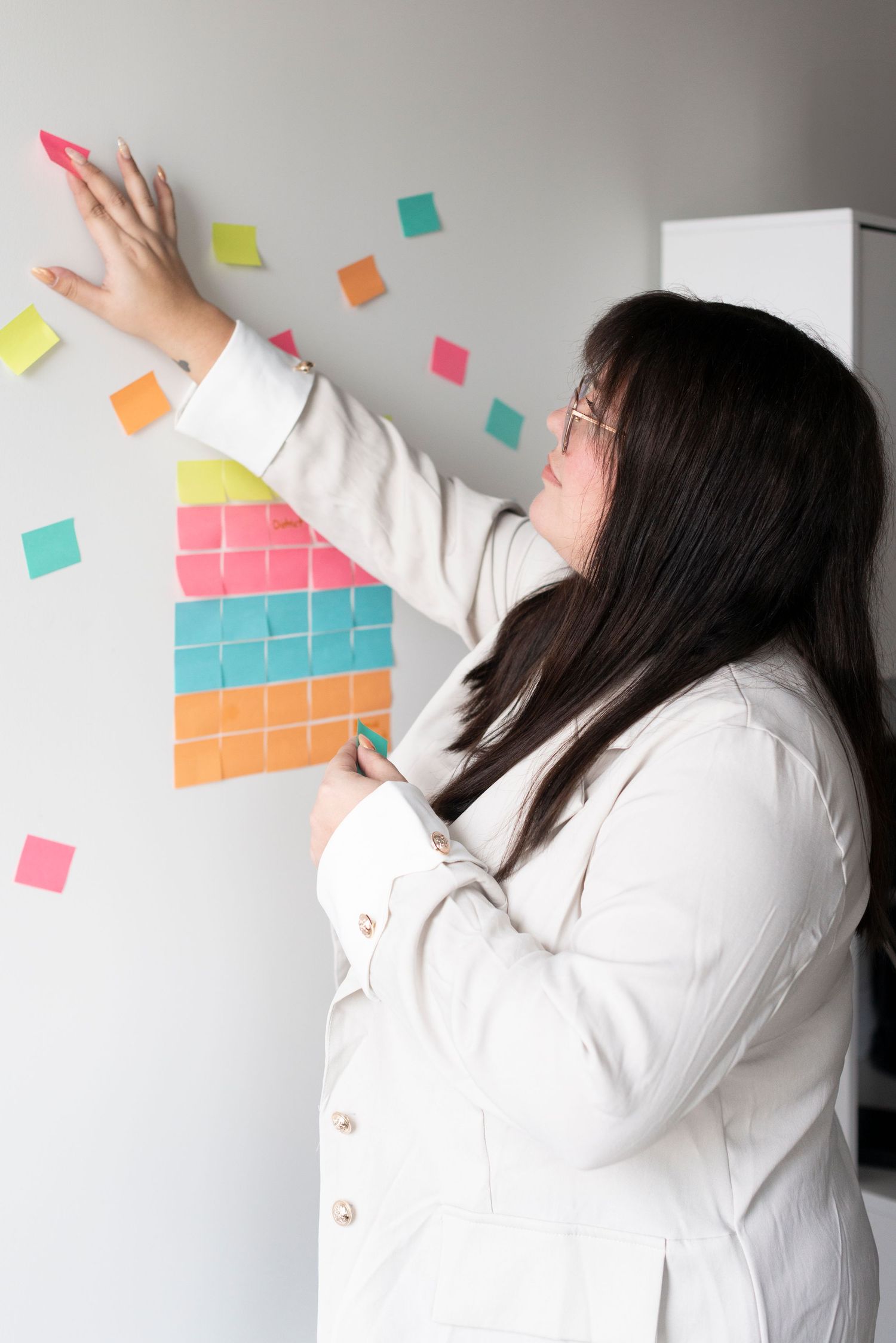 Person in white coat placing colorful sticky notes in a pattern on a white wall.